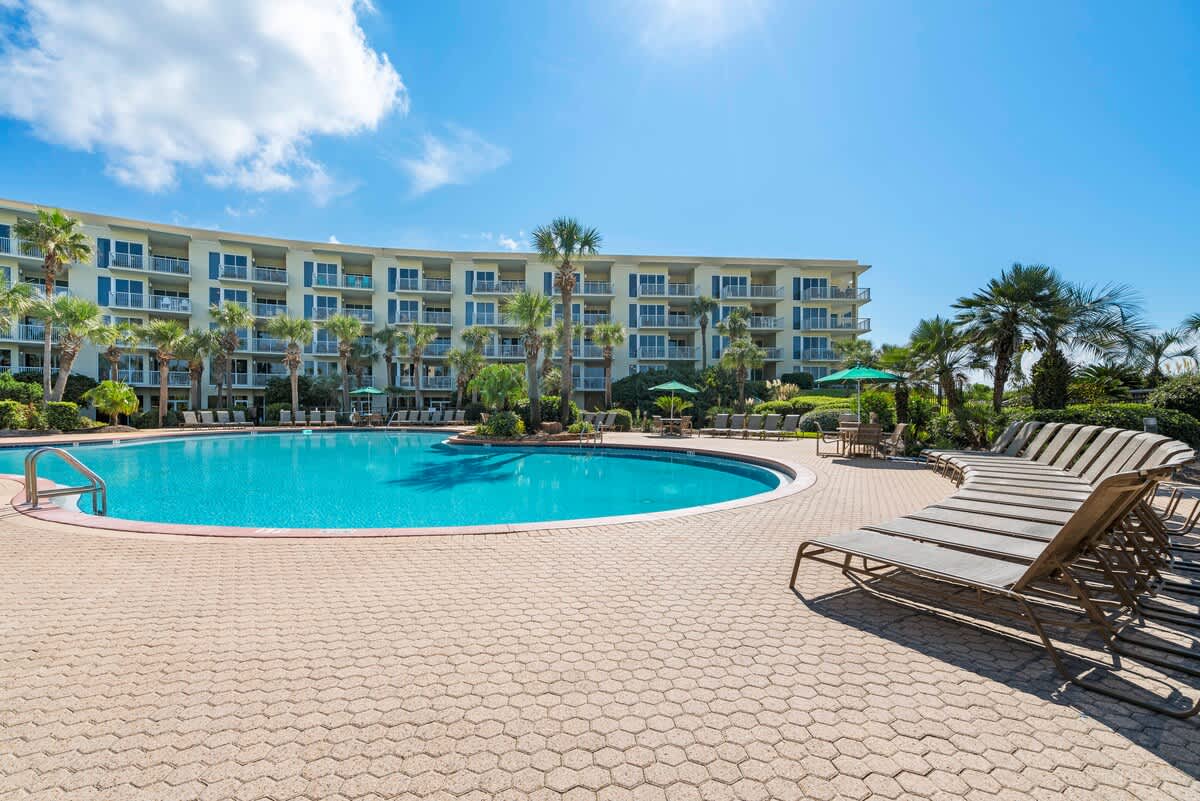 An inviting outdoor pool area featuring a large swimming pool, numerous lounge chairs arranged on a paver deck, and lush palm trees, with a multi-story resort building in the background.