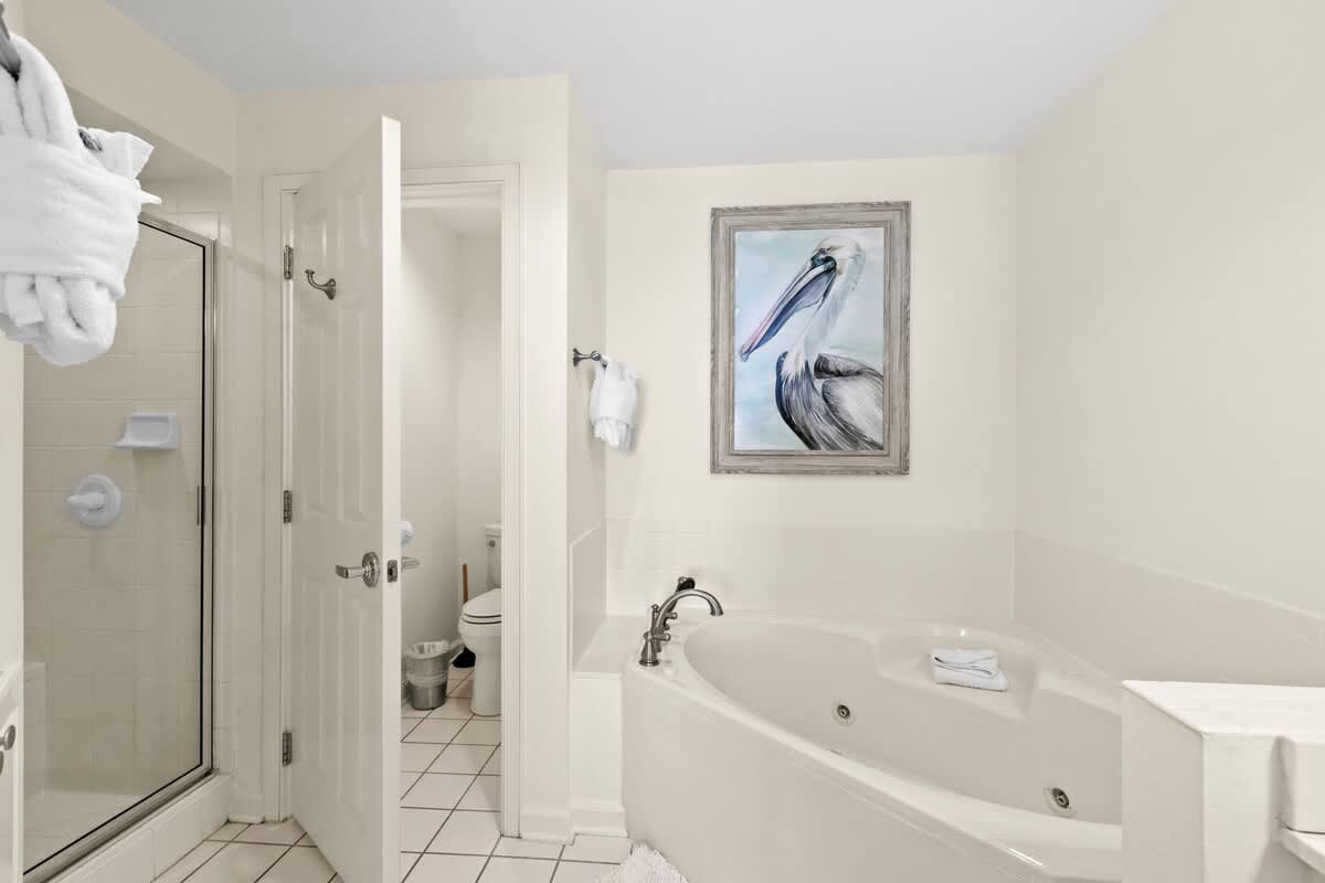 View of the en-suite bathroom showing the corner jetted tub, a separate shower stall, and a doorway to the toilet area, with a pelican painting above the tub.