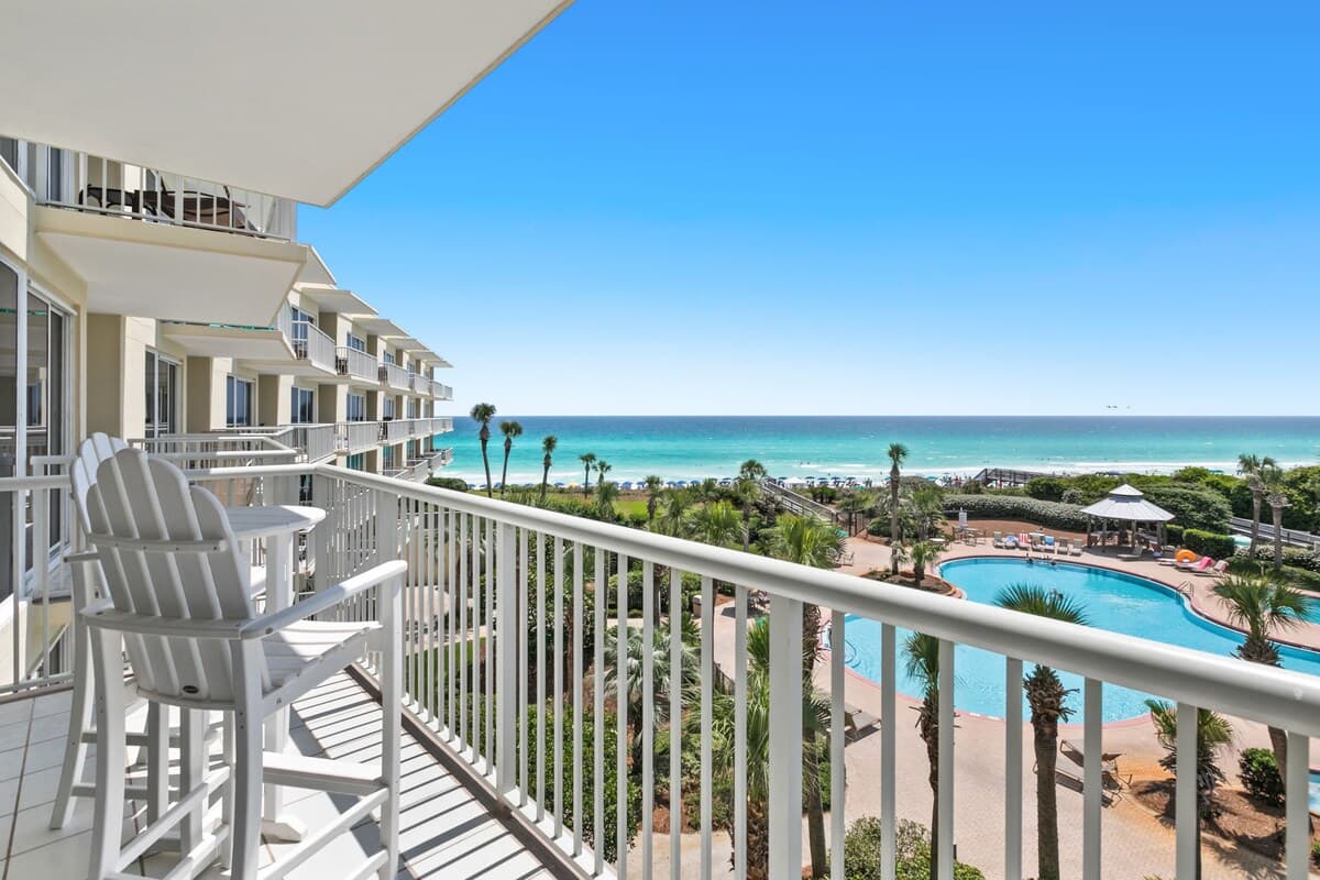 A balcony with stunning views of a large resort pool, lush palm trees, and the sparkling ocean beyond, featuring white Adirondack-style high-top chairs and a small table.