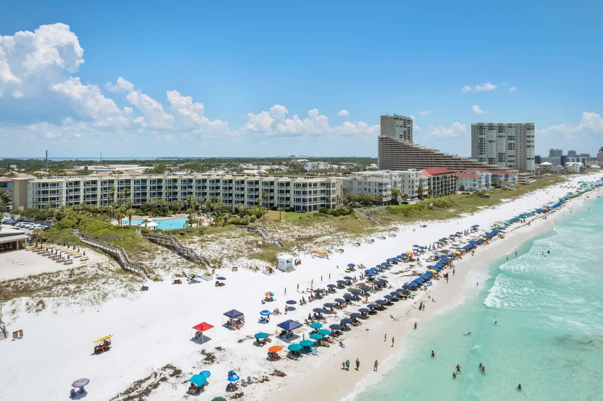 Expansive aerial view of the long, white sand beach lined with countless beach umbrellas and people enjoying the turquoise ocean, with the condominium property and other buildings visible further inland under a partly cloudy blue sky.