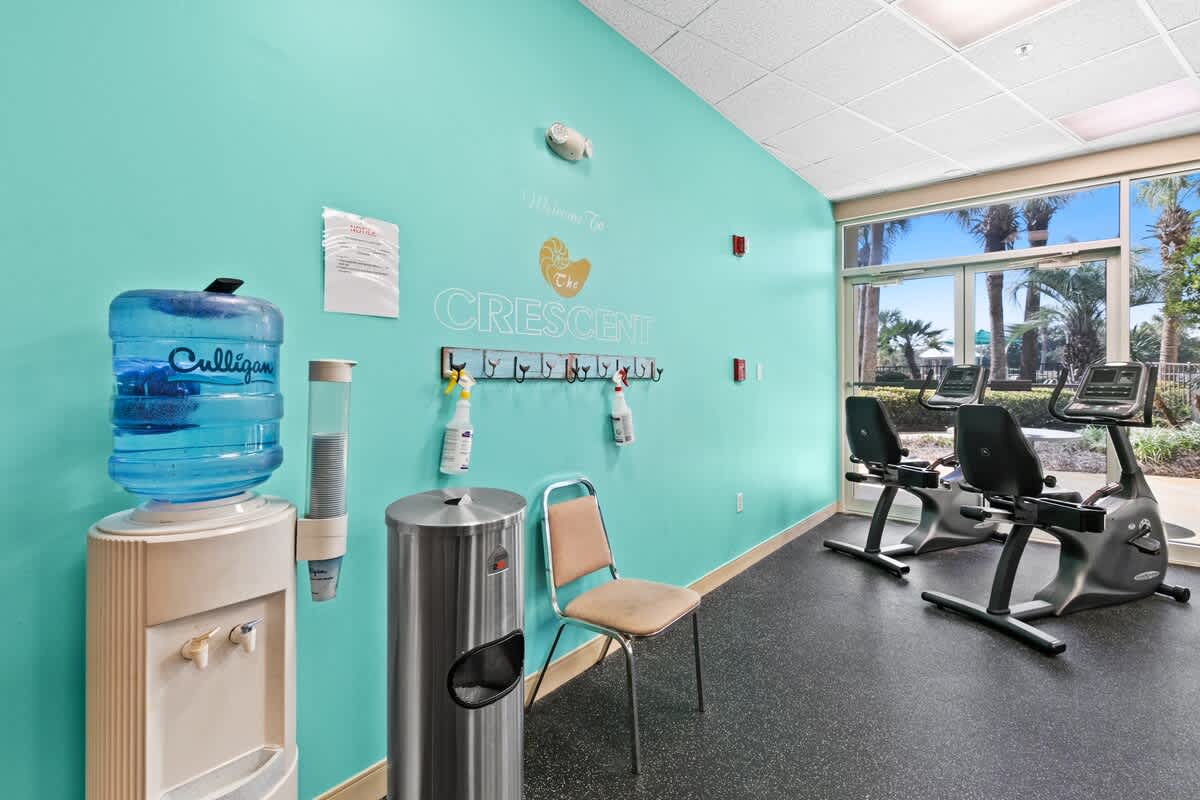 A section of the fitness room with a vibrant teal accent wall featuring a "Welcome To The Crescent" sign, a water cooler, a trash can, and a chair. Two stationary bikes are visible near large windows overlooking palm trees.