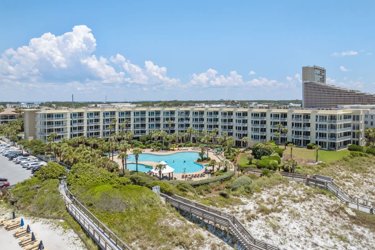 Aerial perspective of the multi-story condominium building and its large, freeform swimming pool, surrounded by palm trees and green spaces, with a wooden boardwalk winding through sandy dunes towards the distant white sand beach.