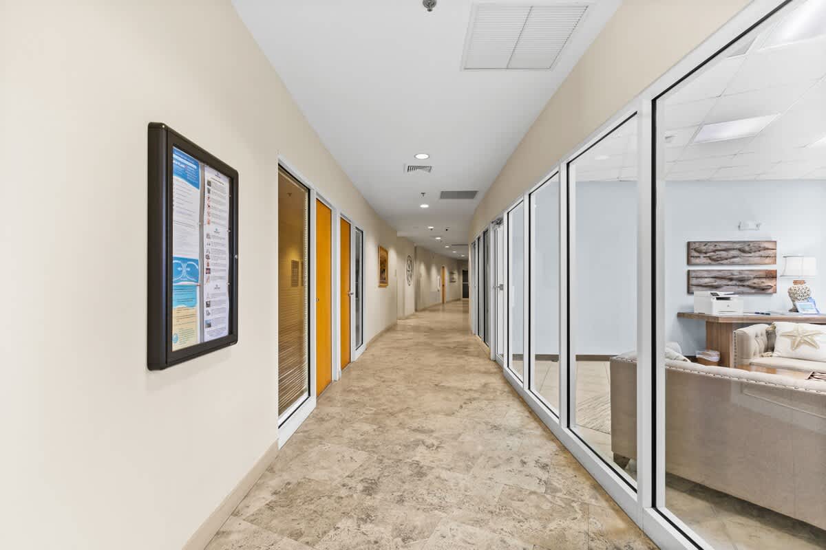 A long, brightly lit hallway with light tiled flooring and a drop ceiling, featuring a series of glass-paneled rooms or offices along one side. A bulletin board with information is mounted on the opposite wall.
