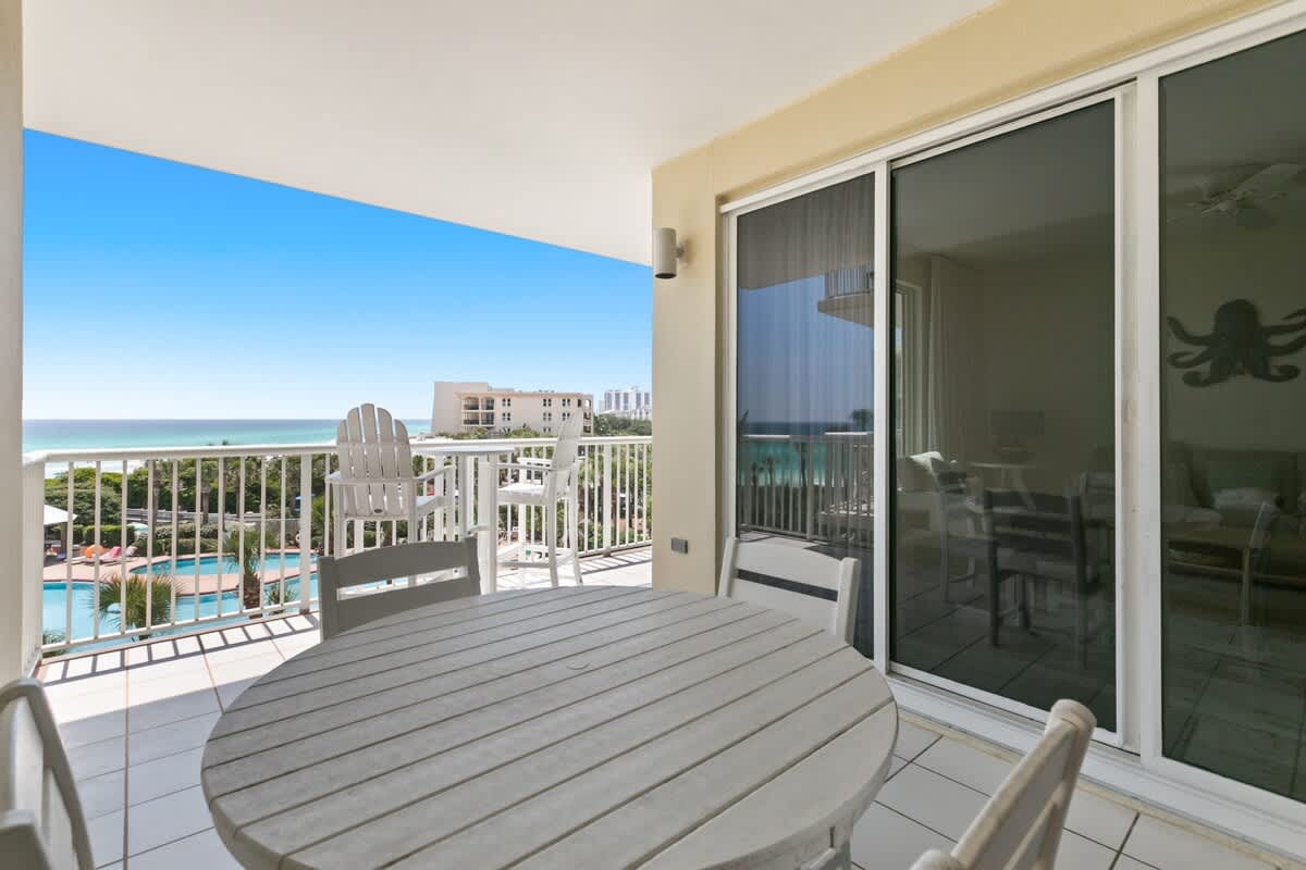 A spacious balcony offering panoramic ocean and pool views, furnished with a round white outdoor dining table and chairs, and featuring a white metal railing.