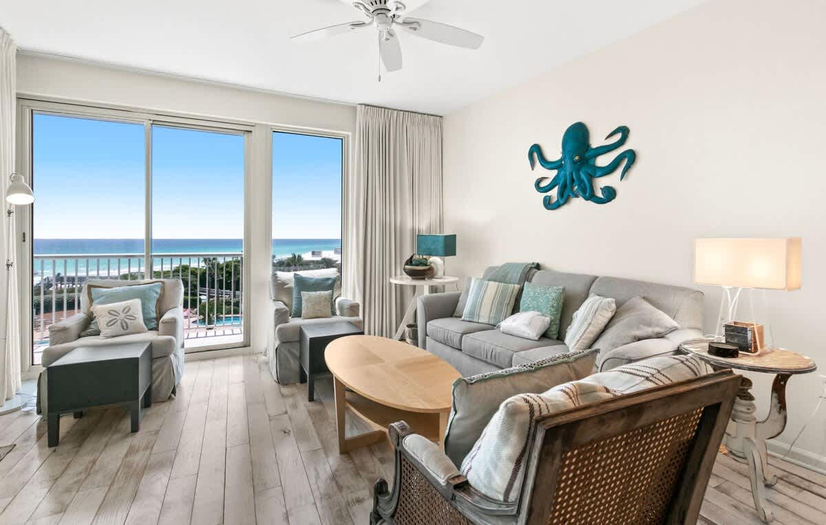 Coastal-themed living room featuring a grey sofa with accent pillows, two armchairs, and a distinctive blue octopus wall art, all overlooking the ocean through large glass doors.