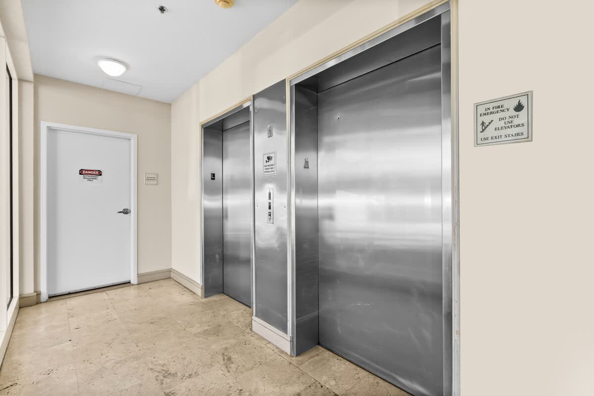 A well-lit common hallway featuring two stainless steel elevators and a white door with a 'DANGER' sign, all set against light-colored walls and a tiled floor.