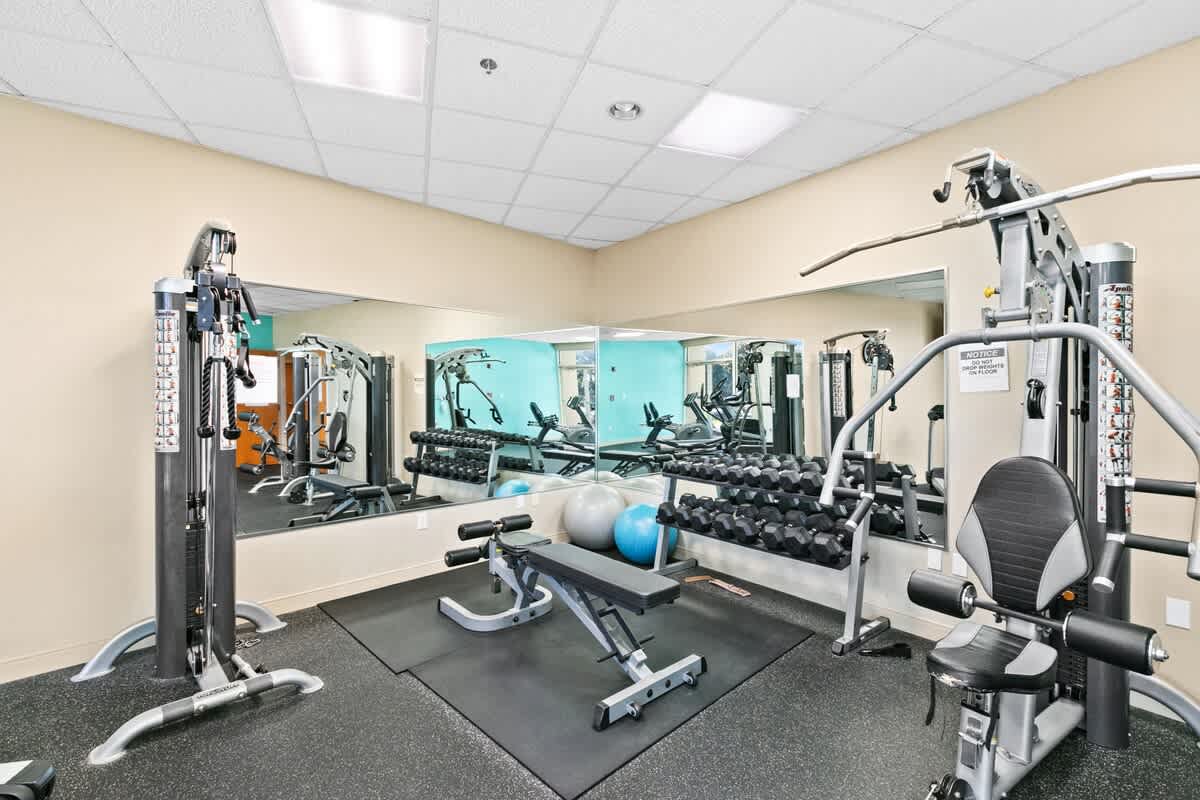 A section of the communal fitness center featuring weight machines, a rack of dumbbells, an adjustable exercise bench, and large wall mirrors reflecting the equipment.