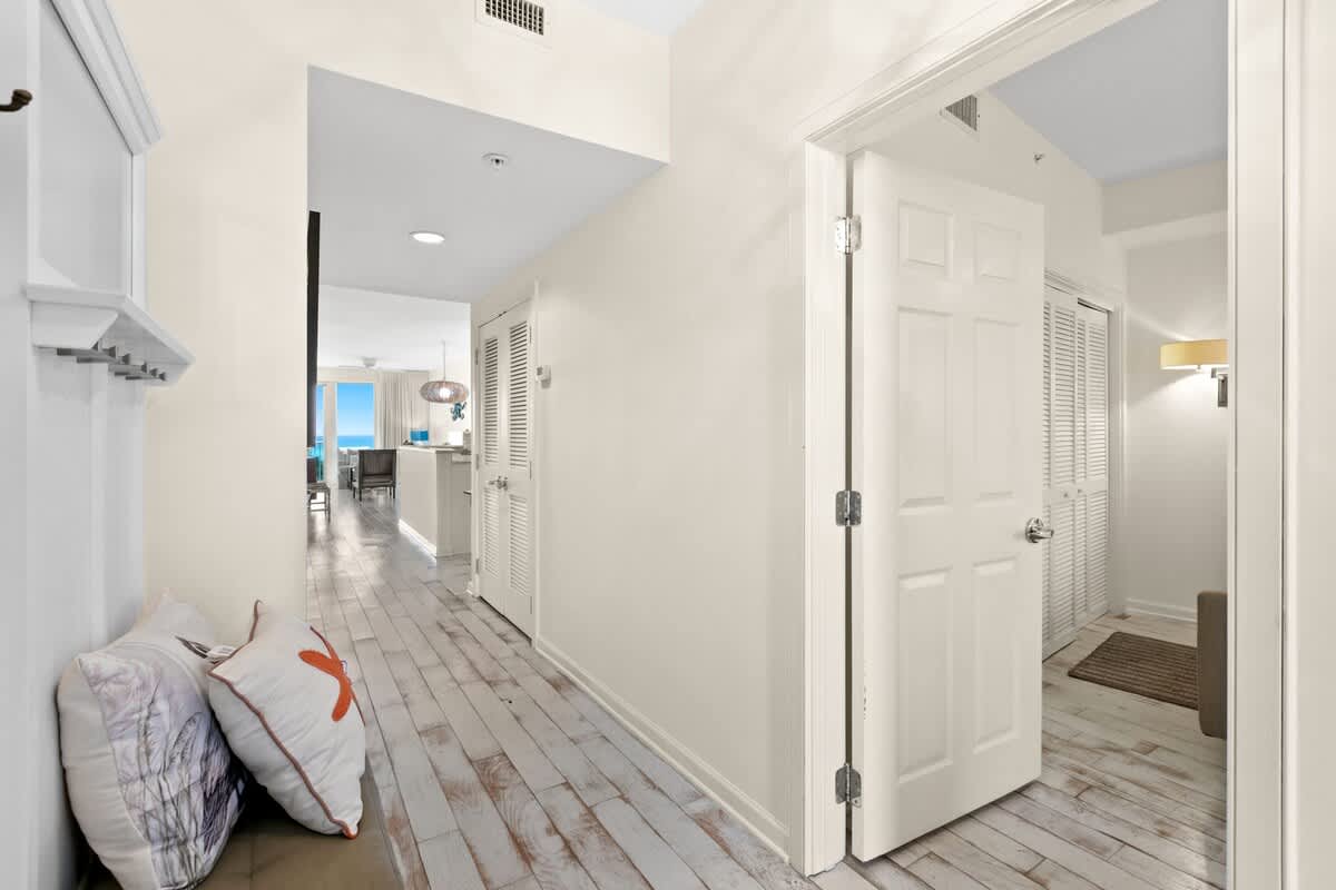 A bright foyer with light, distressed wood-look flooring and white walls, leading into the main living area with an open doorway to another room. Decorative pillows rest on a bench to the left.