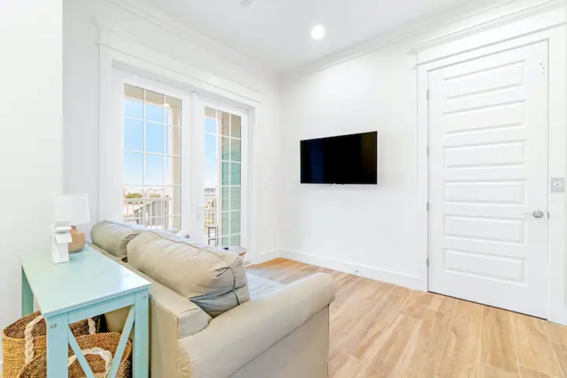 Cozy living area featuring a light-colored sofa facing a wall-mounted television. A light blue console table with a woven basket sits beside the sofa, and double glass doors open to a balcony, providing natural light.