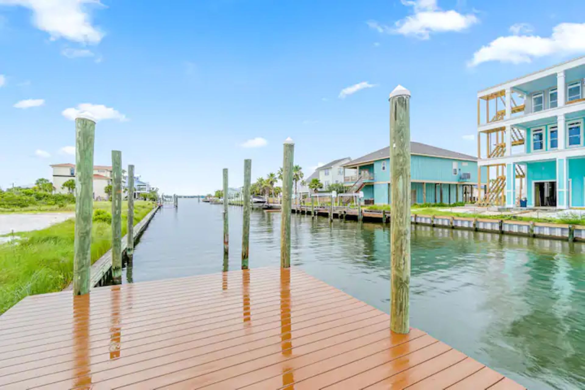 A wooden dock extending into a calm canal, lined with green pilings and surrounded by lush green grass, with a light blue multi-story house visible in the background.