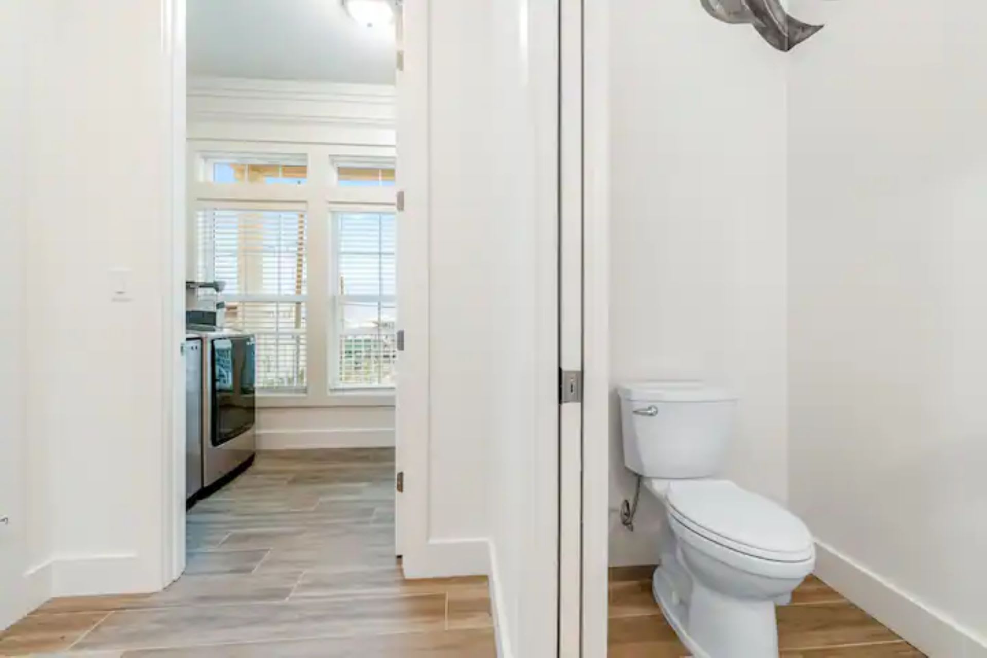 Narrow hallway featuring a discreet half-bathroom with a white toilet. Through an open doorway, a modern kitchen with stainless steel appliances and windows with blinds is visible, suggesting an open layout.