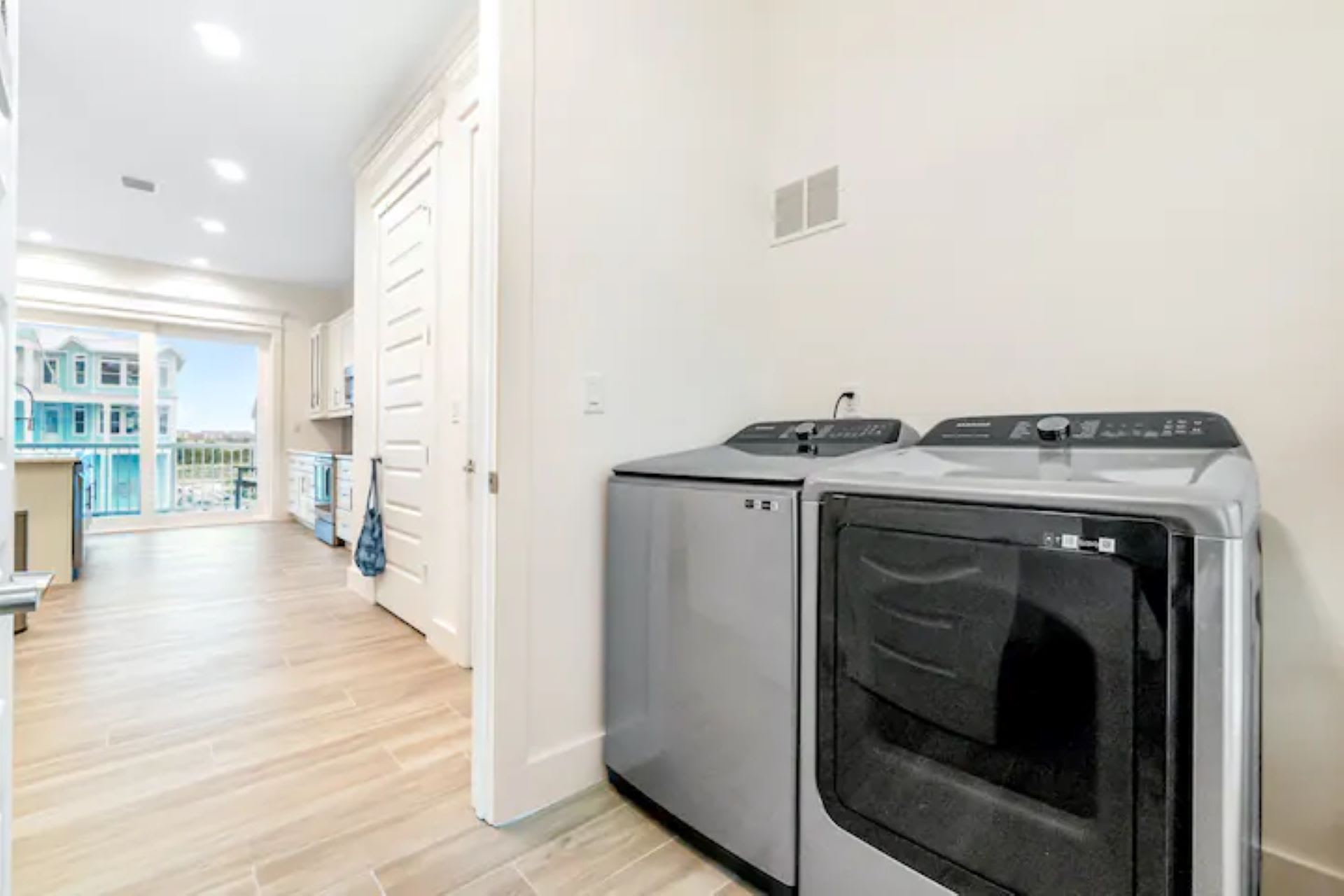 Dedicated laundry area in a hallway with a silver washer and dryer, featuring wood-look tile flooring and a view towards a kitchen or living area.