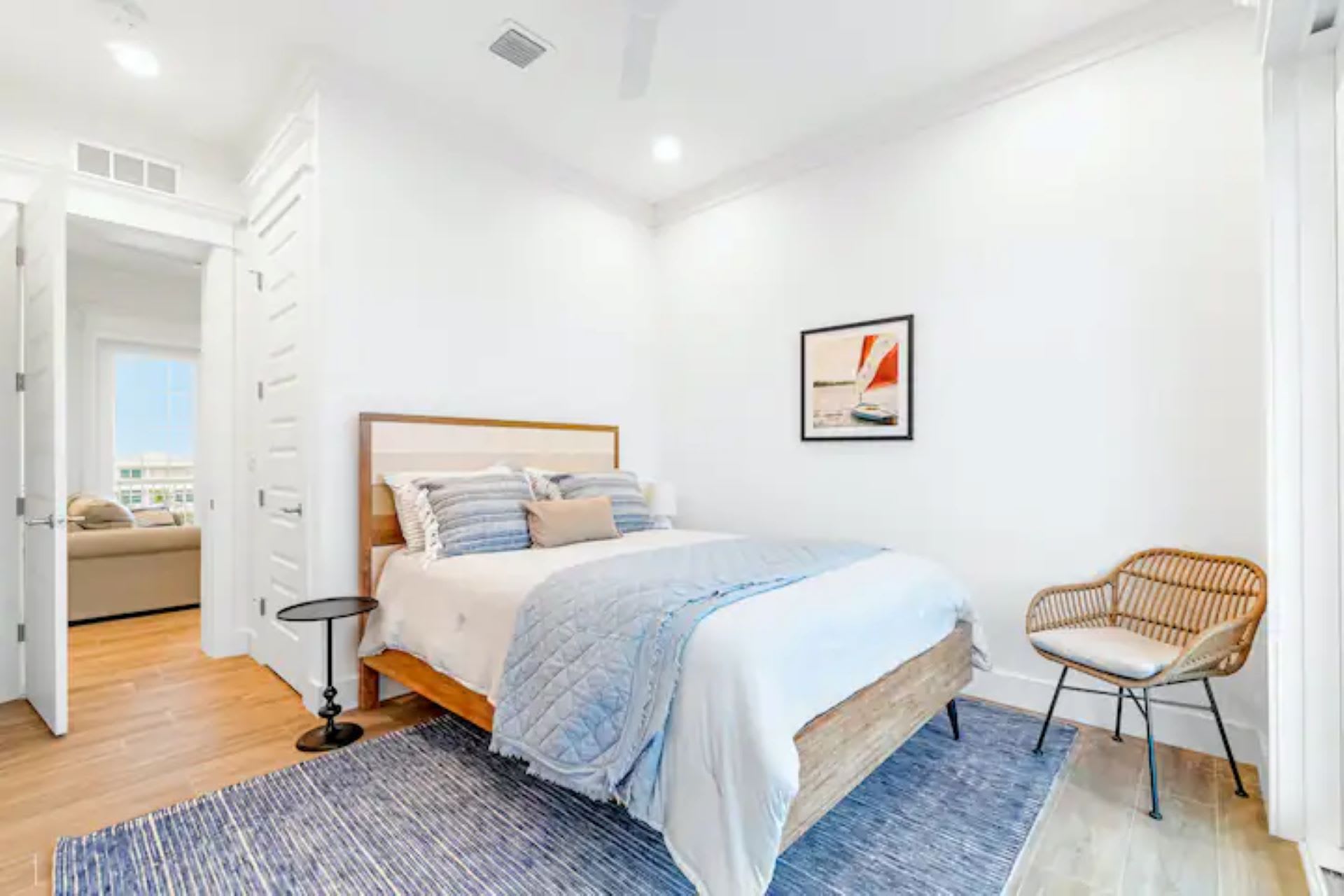 Inviting bedroom with a queen-sized bed featuring a wooden headboard and blue and white striped bedding. A small round black nightstand and a wicker chair flank the bed, while a blue patterned rug adds color to the light wood floors.