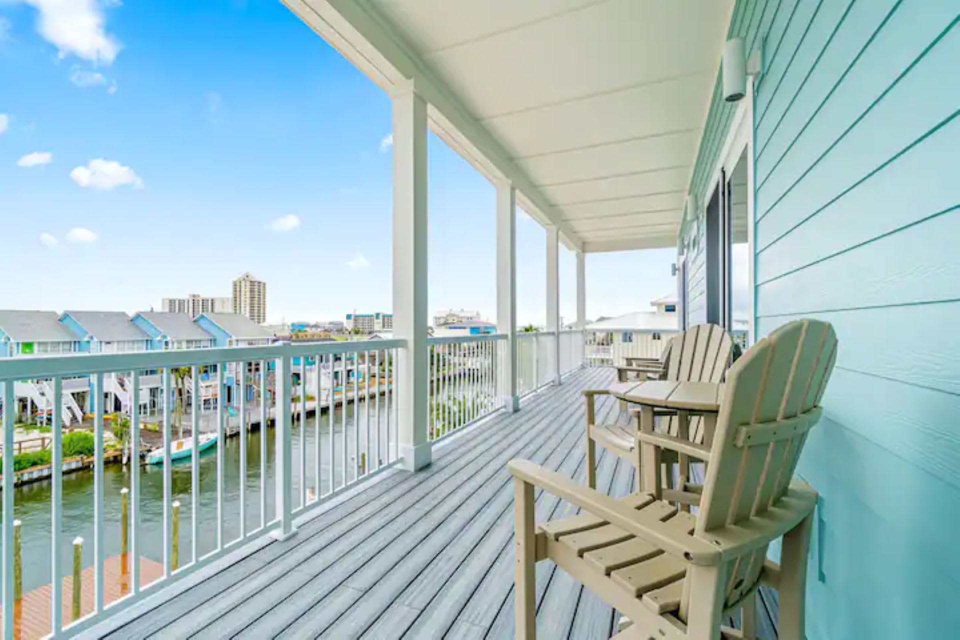 A spacious covered deck with light blue siding and grey plank flooring, furnished with two light-colored Adirondack chairs and a small table, offering views of a canal and distant buildings.