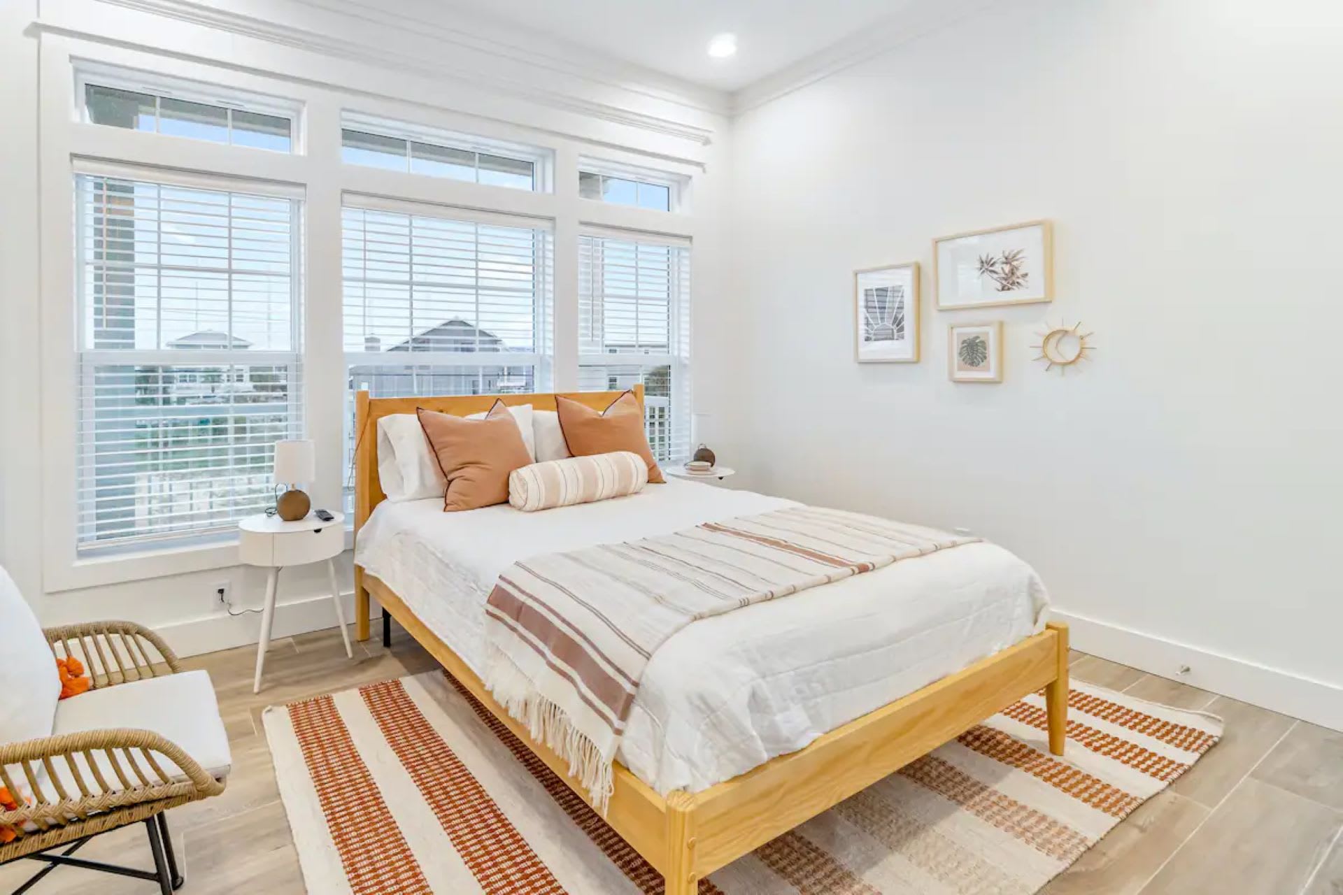 Serene bedroom featuring a queen-sized bed with a light wood frame and white bedding accented with warm orange and brown tones. Large windows with blinds offer ample natural light, complemented by framed artwork and a wicker chair.