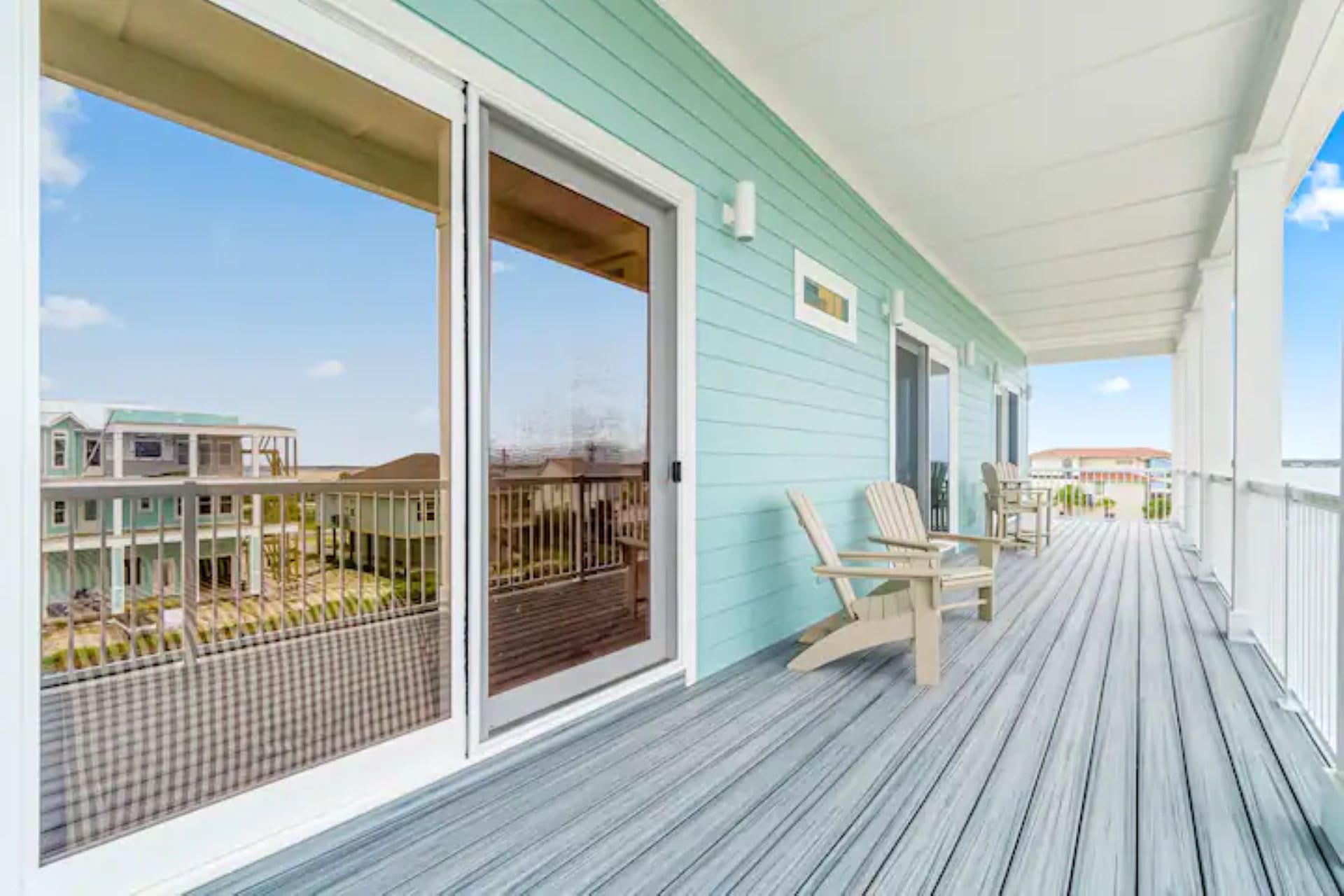 A long, covered deck with light blue siding and grey plank flooring, featuring two light-colored Adirondack chairs and large sliding glass doors reflecting the sky and distant houses.