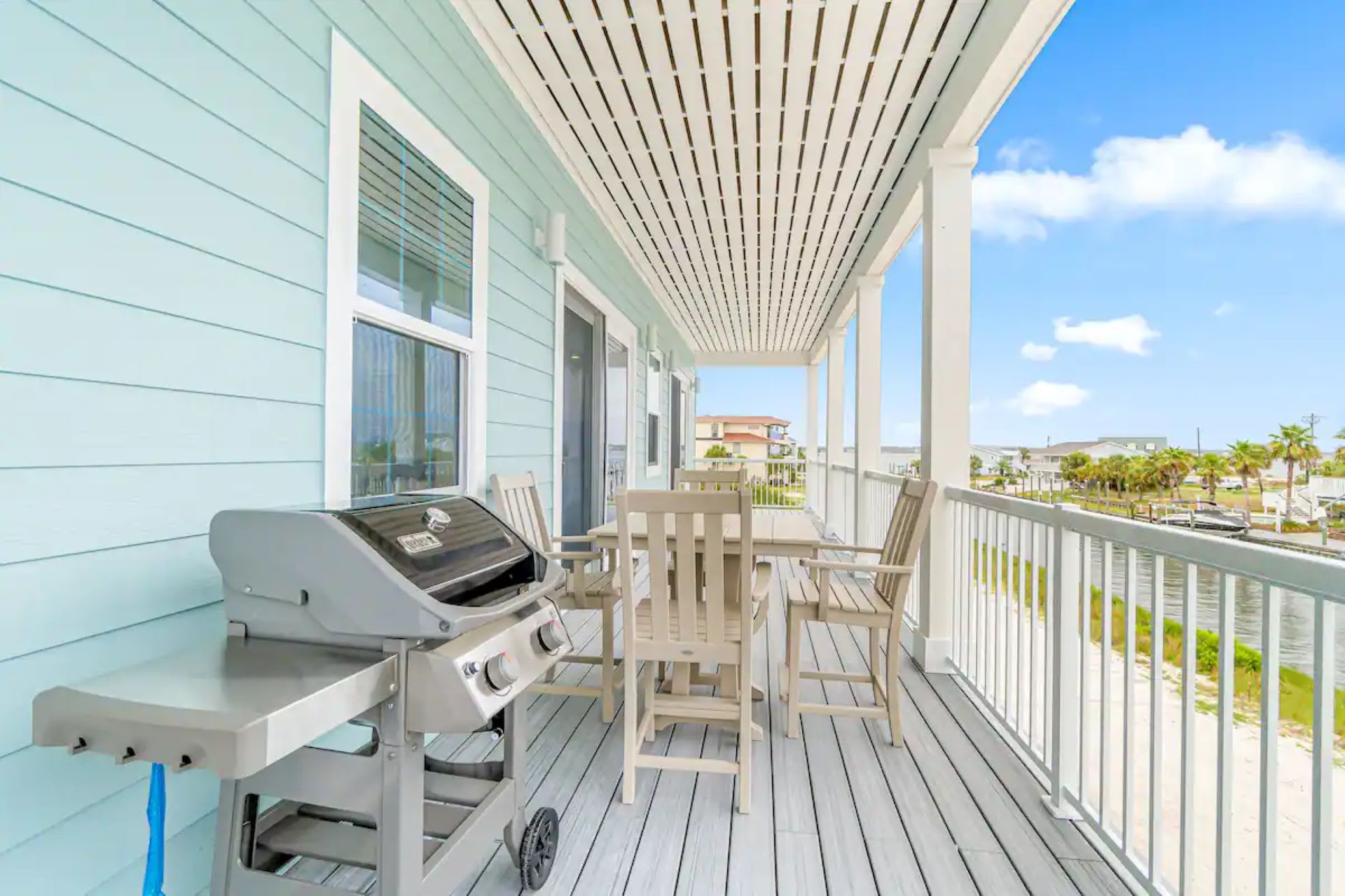 A covered outdoor deck with light blue siding and grey plank flooring, equipped with a stainless steel BBQ grill and a wooden outdoor dining set, offering views of a canal.
