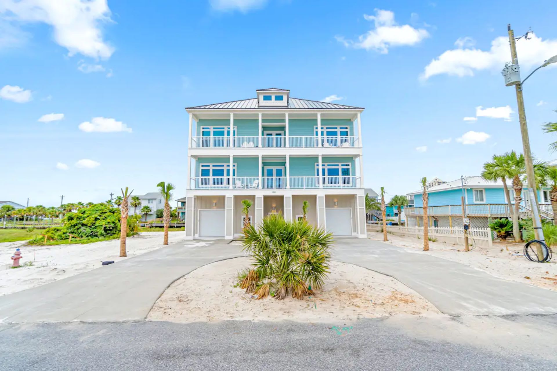 Grand three-story vacation home with light blue siding and multiple white-railed balconies on each level. The ground floor features garages, and a circular driveway leads up to the entrance, surrounded by palm trees and sandy landscaping.