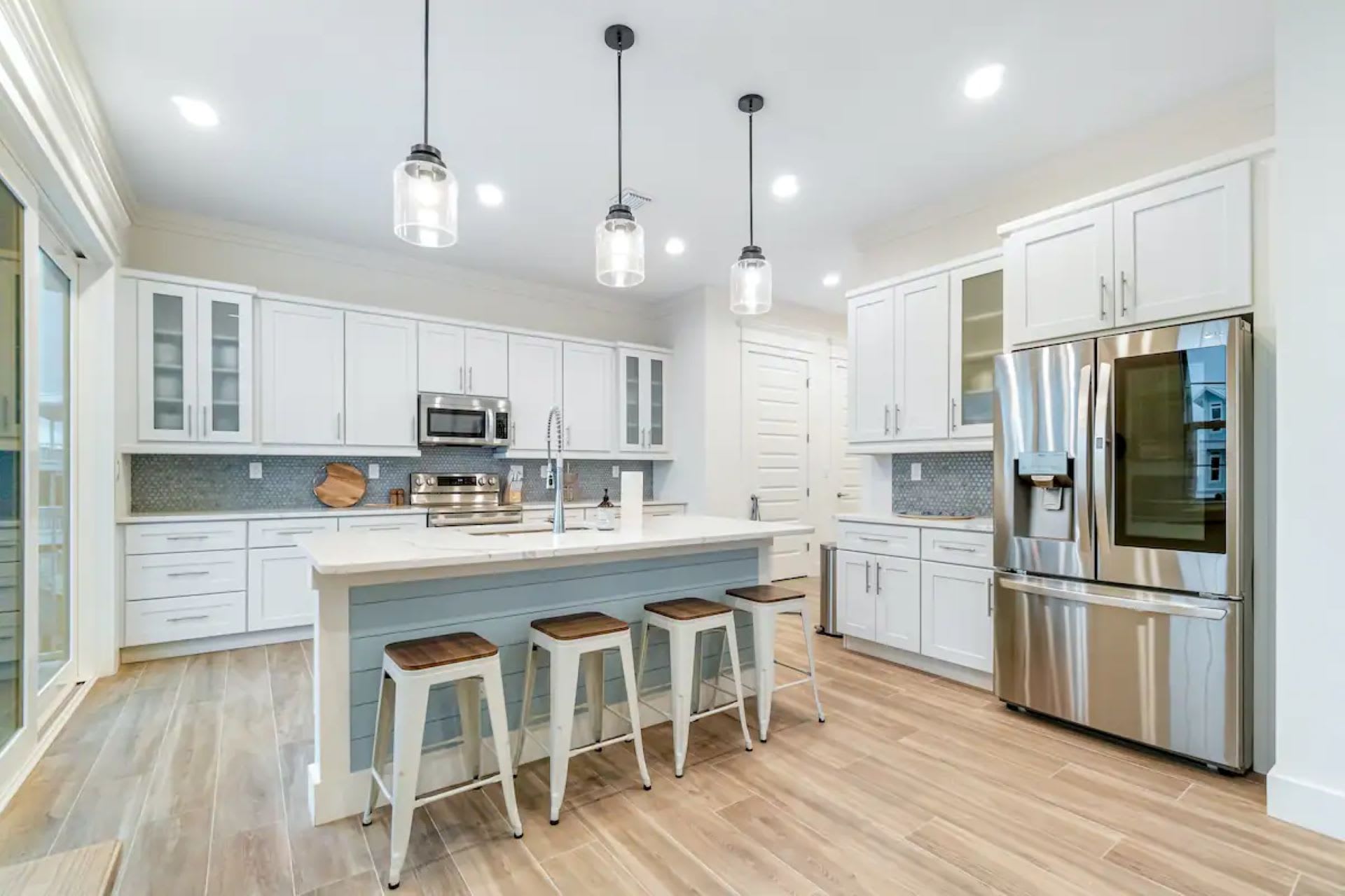 The bright kitchen showcases white cabinetry, a large stainless steel French door refrigerator, a light blue island with seating, and glass-front upper cabinets, all under recessed lighting.