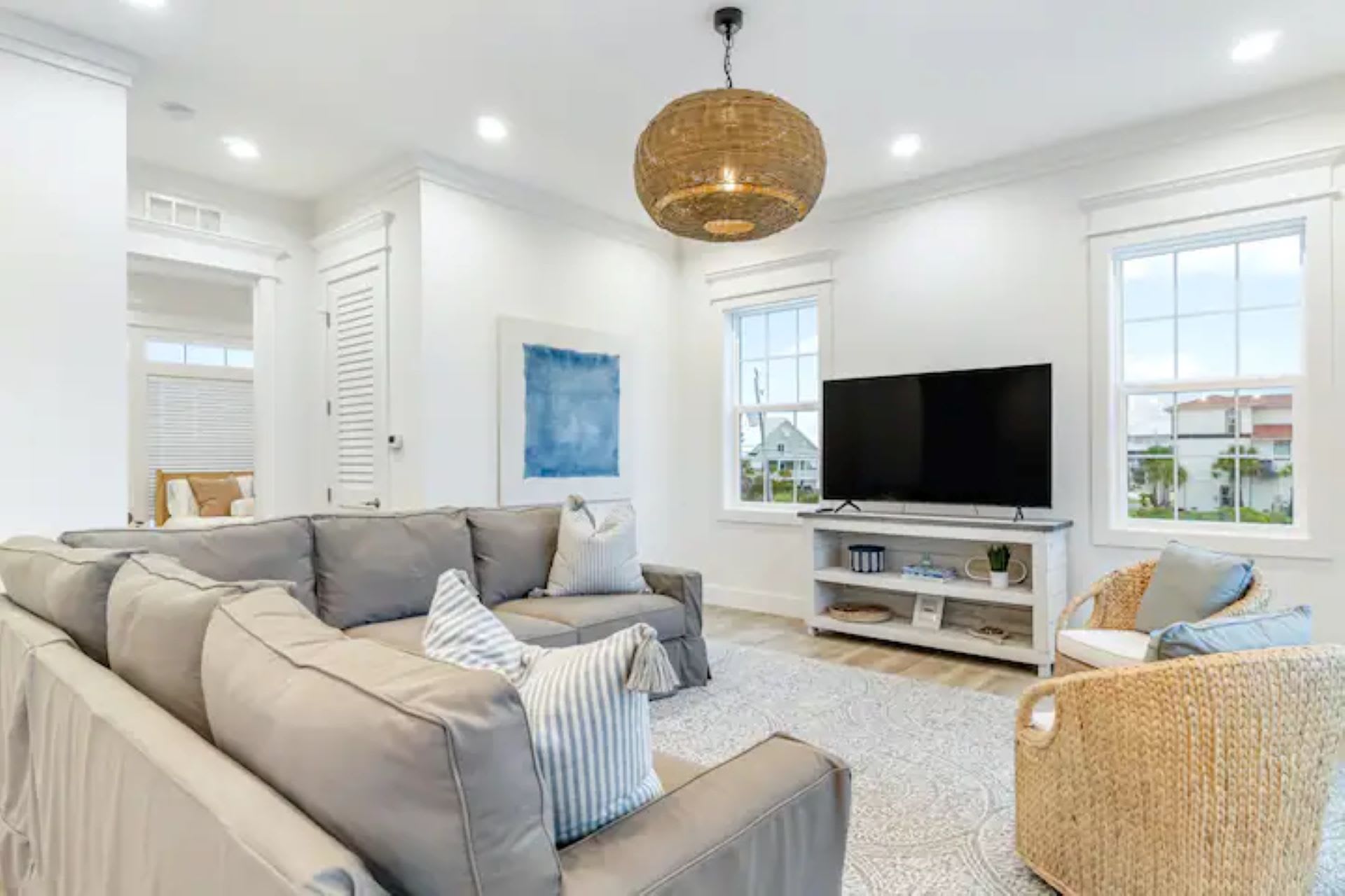 The living room includes a large wall-mounted TV on a white console, a gray sectional sofa, and two large windows with white blinds, offering views of neighboring houses.