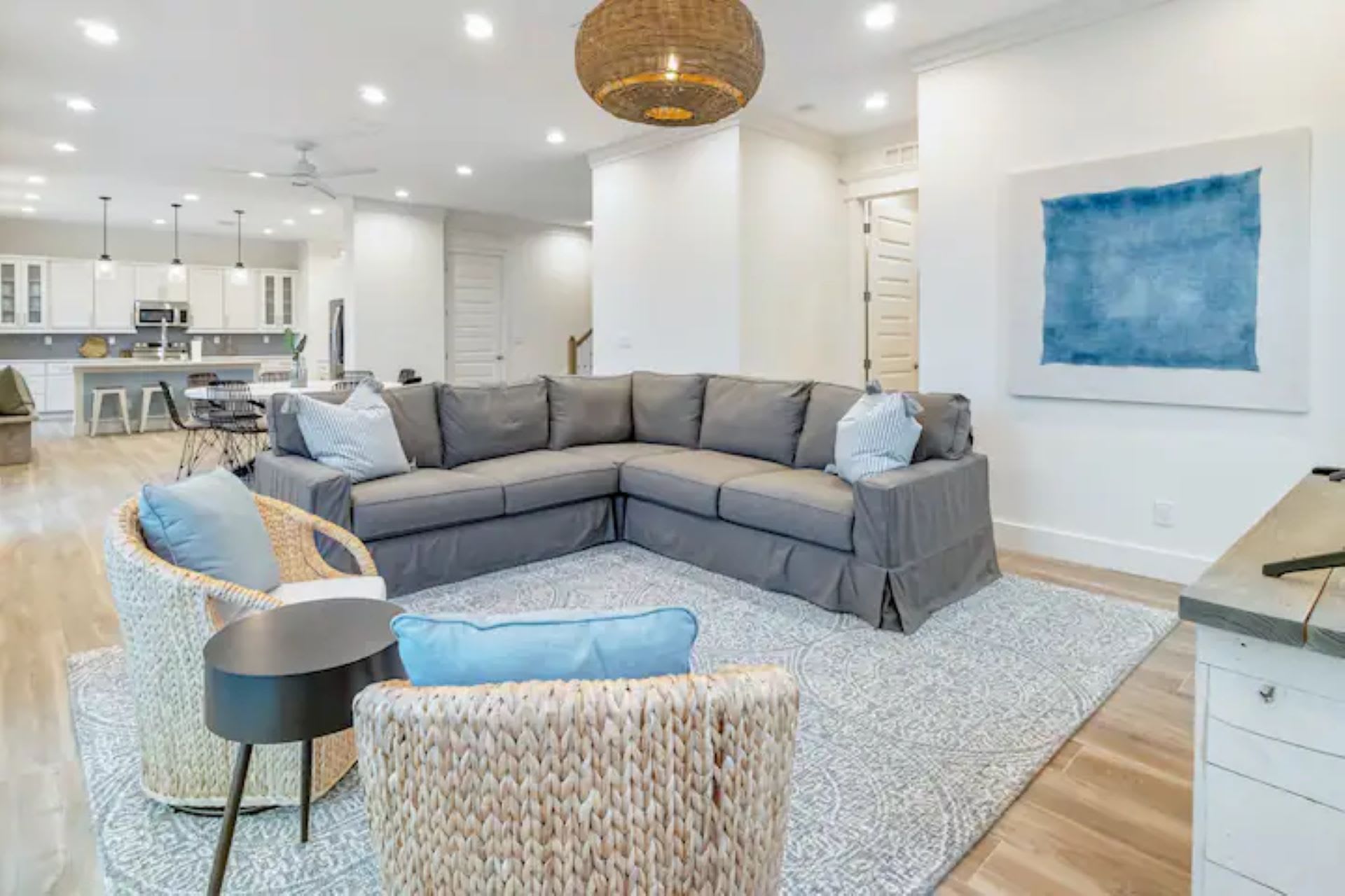 A spacious living room features a large gray sectional sofa, two woven armchairs, a patterned rug, and a unique woven pendant light, with a large blue abstract painting on the wall.