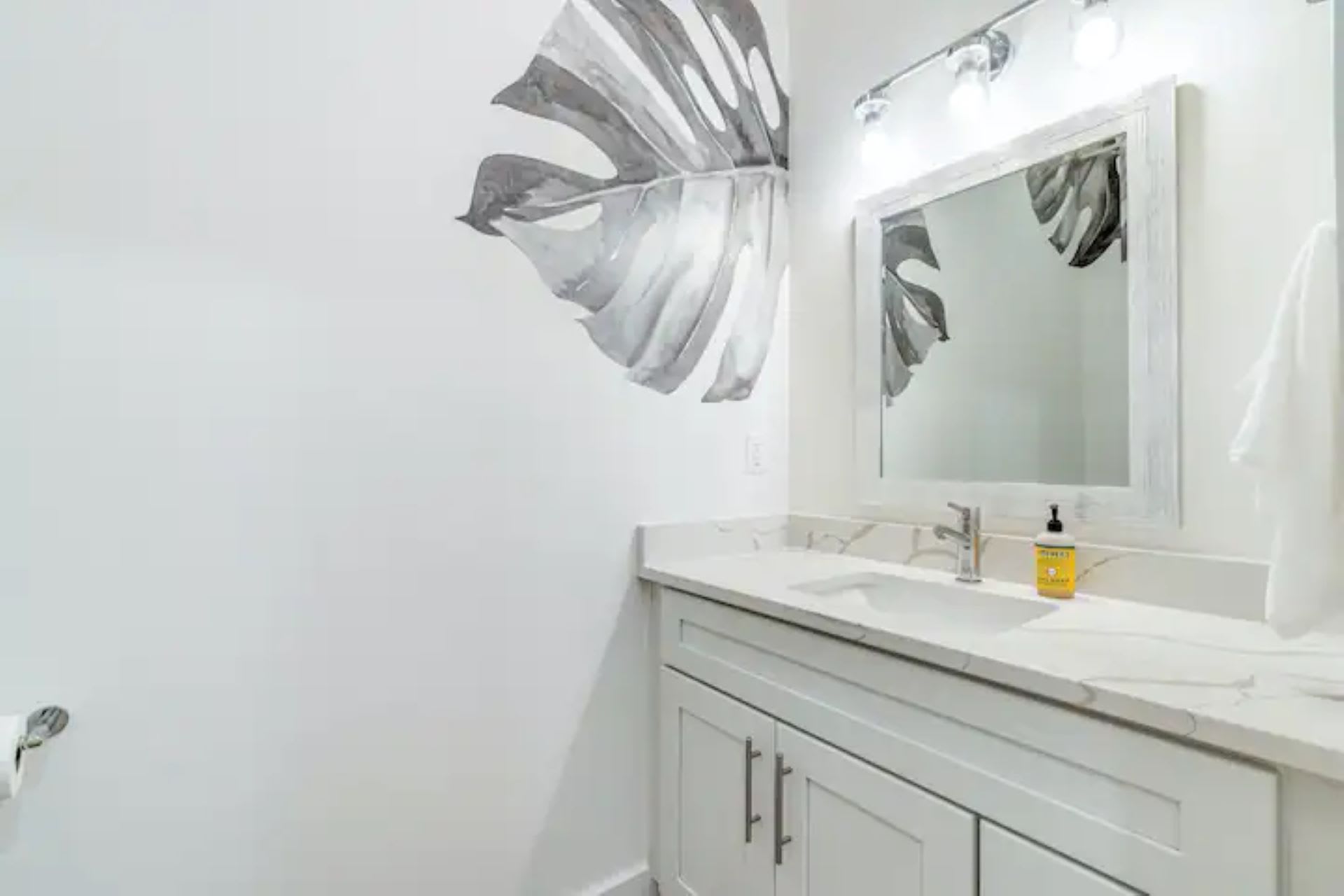 Compact half bathroom with a single vanity, white countertop, framed mirror, and a large grey leaf decal on the white wall.