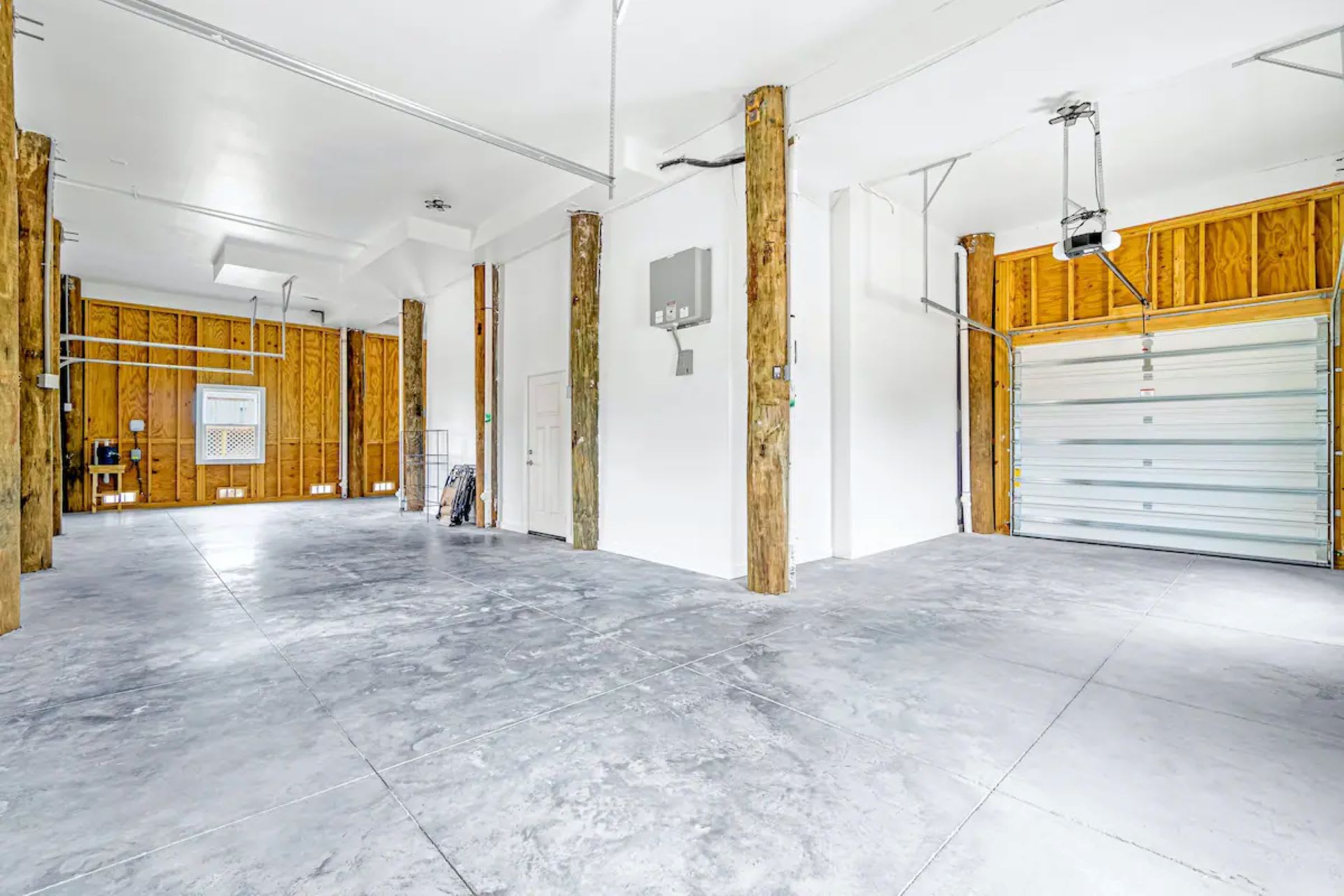A spacious, open garage area features concrete floors, exposed wooden support beams, and two large garage doors, providing ample parking and storage space under the elevated house.