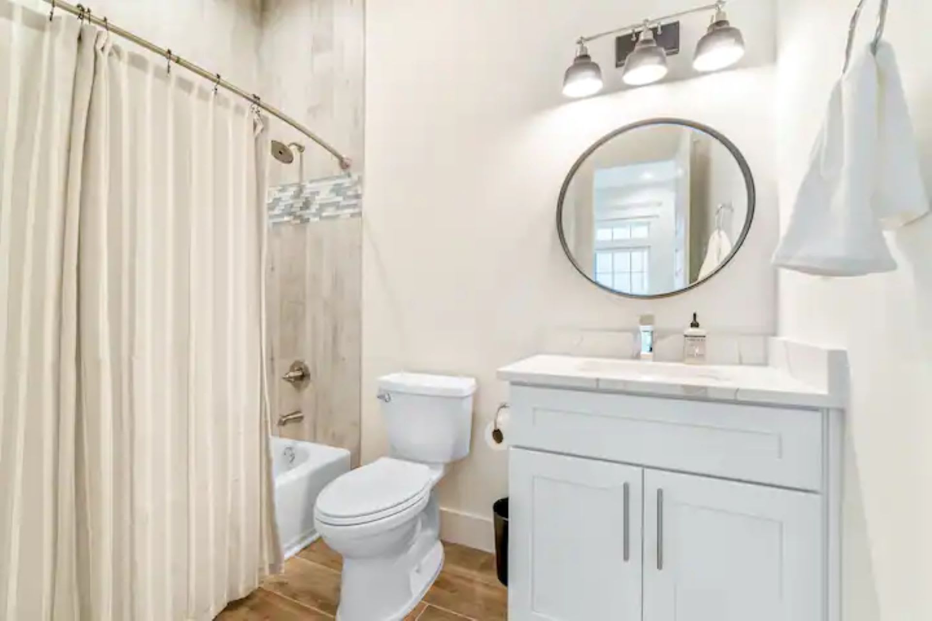 Compact bathroom with a white bathtub and shower, enclosed by a striped shower curtain. It includes a white toilet, a single vanity with a white countertop, and a round mirror illuminated by a three-light fixture.