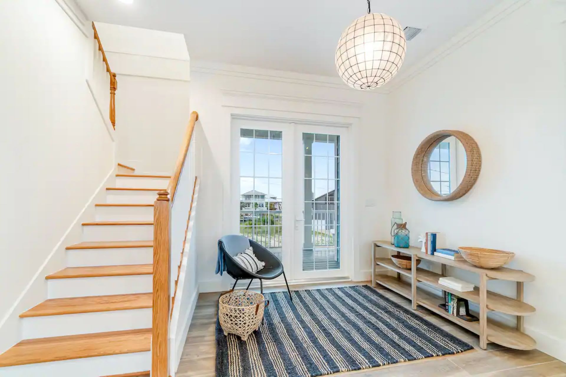 Inviting foyer featuring a wooden staircase with white risers, a blue and white striped rug, and a light wood console table with decorative items. A round mirror hangs above the console, and double glass doors lead to the exterior.