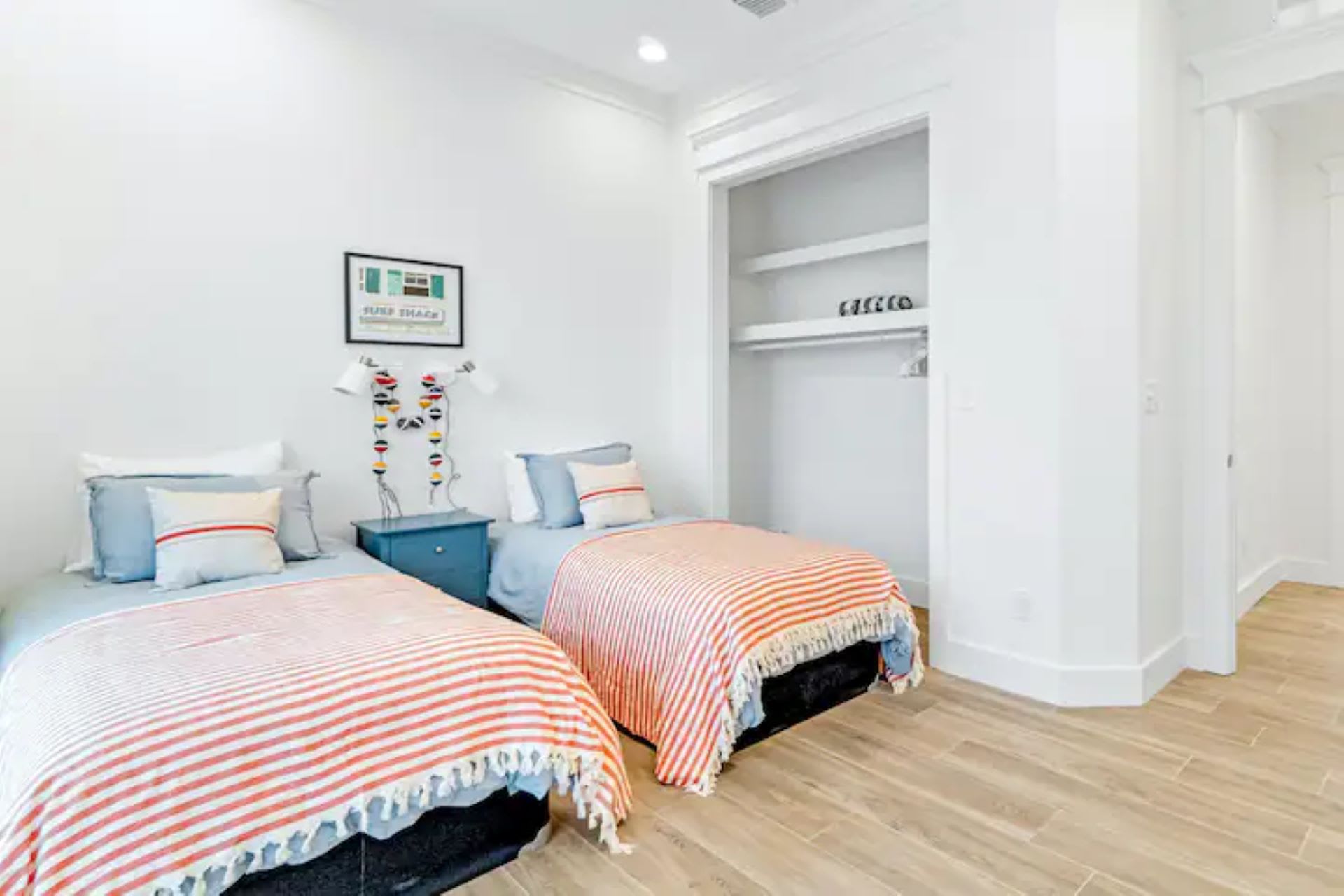 Kids' bedroom with two twin beds featuring colorful striped blankets, a blue nightstand, and an open closet with shelves, all on wood-look tile flooring.