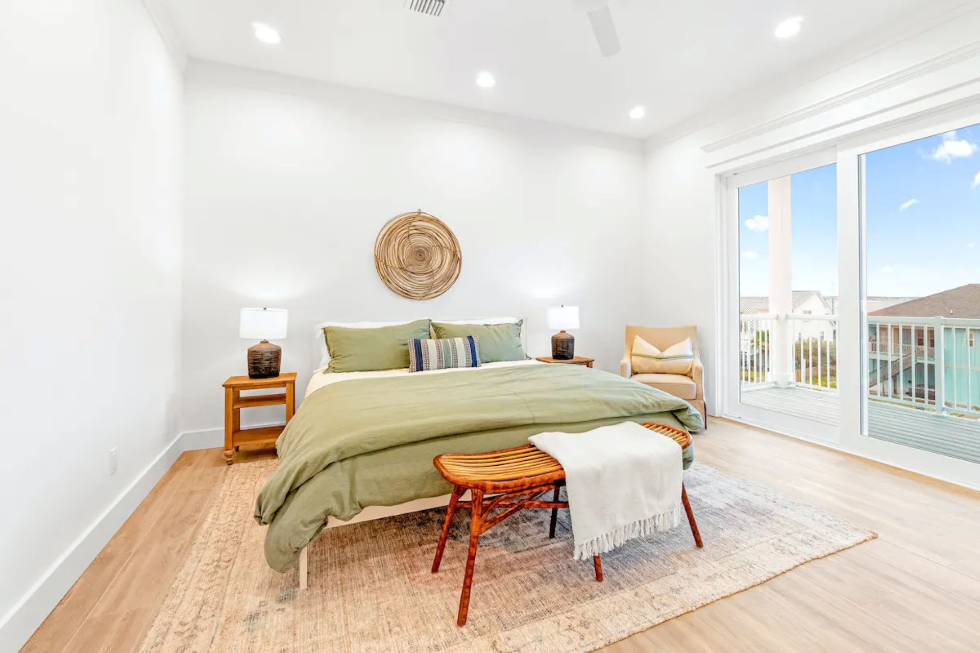 Bright bedroom with a king-sized bed featuring olive green bedding, flanked by wooden nightstands with lamps. A woven wall hanging adorns the white wall, and a large sliding glass door opens to a balcony, offering views of the outdoors.