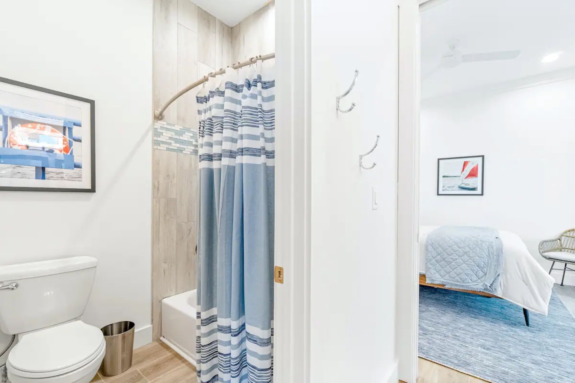 Bright bathroom with a bathtub and shower, enclosed by a blue and white striped shower curtain. A white toilet is visible, and framed artwork hangs on the wall, with a peek into an adjacent bedroom through the open doorway.