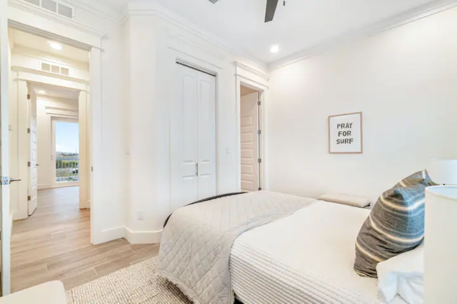 Cozy bedroom with a white quilted bed, a striped accent pillow, and a framed "PRAY FOR SURF" sign on the white wall, featuring wood-look tile flooring.