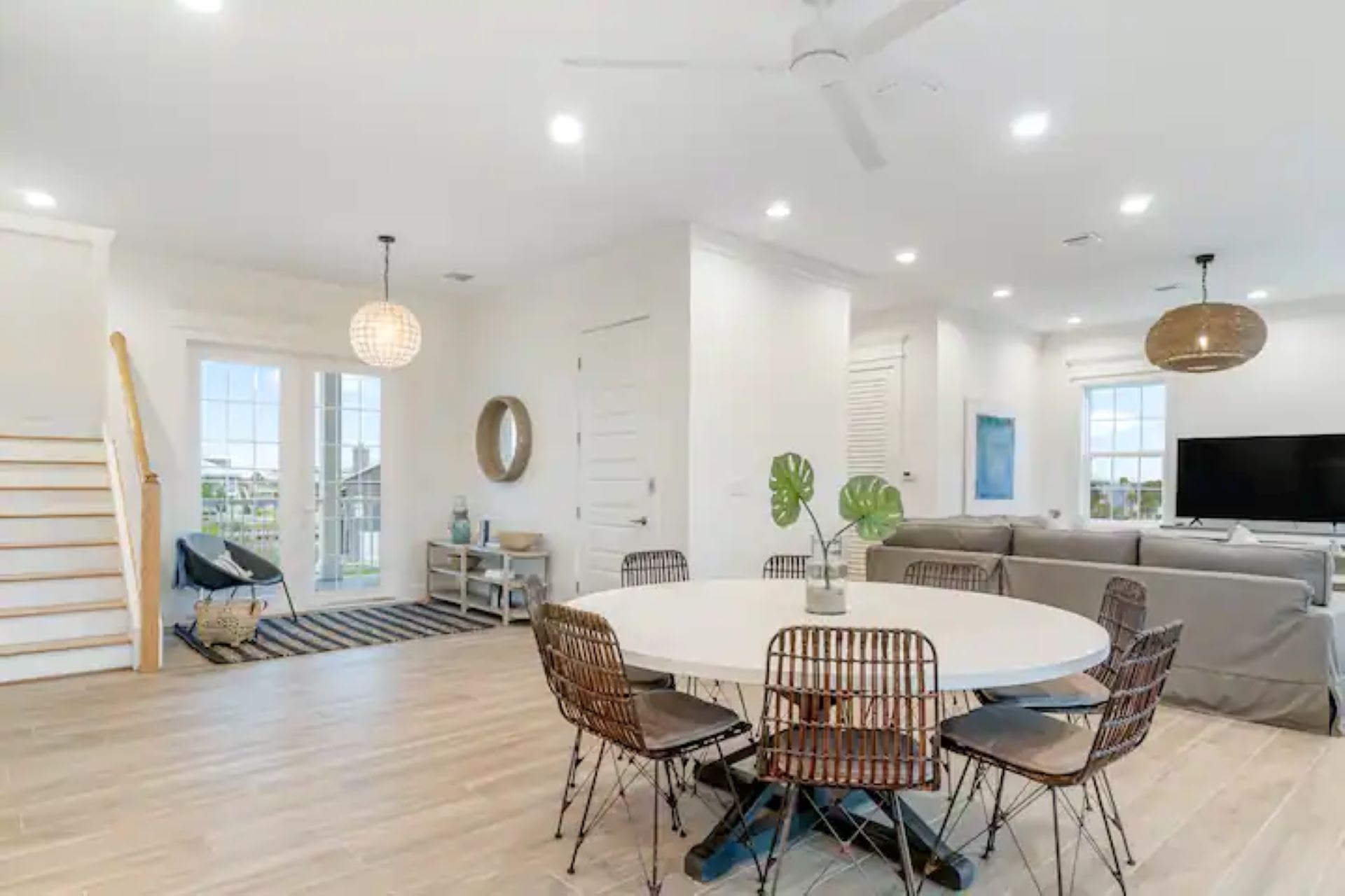 An open-concept dining area features a round white table with six woven chairs, adjacent to the living room, with light wood floors and a staircase leading to an upper level.