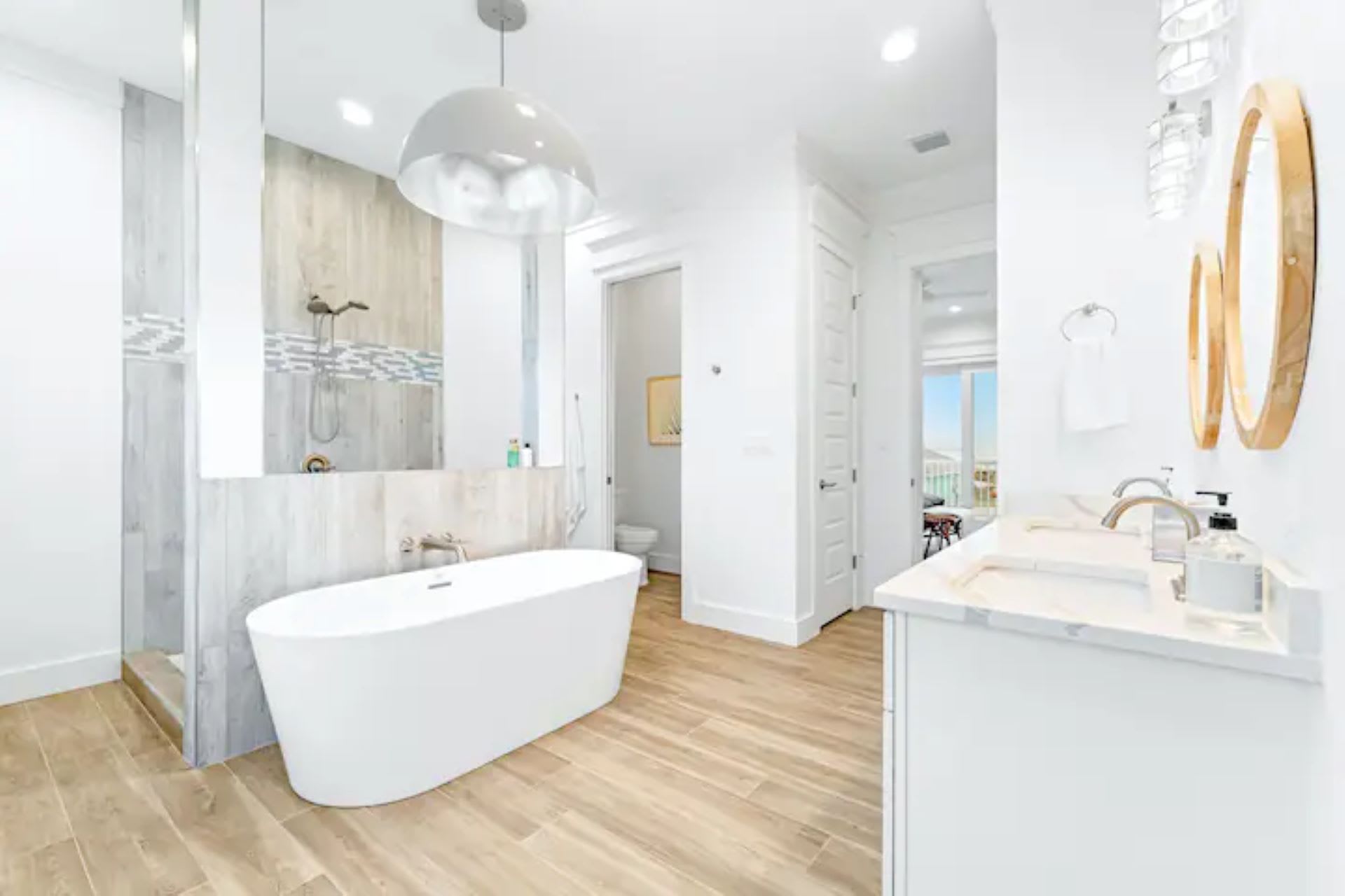 Luxurious master bathroom featuring a freestanding white soaking tub and a spacious walk-in shower with a glass panel. A double vanity with two sinks is illuminated by two oval mirrors and modern pendant lights, all set against light wood-look tile floors.