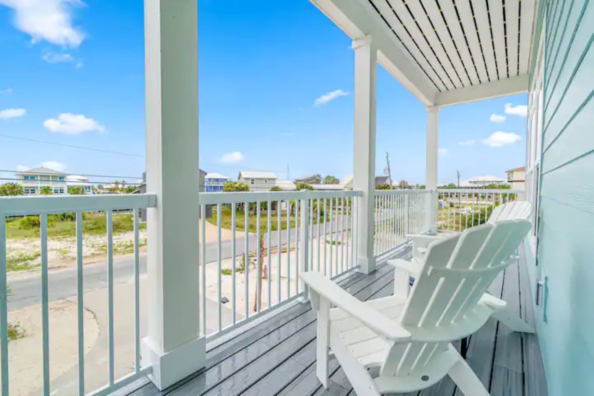 A covered deck with light blue siding and grey plank flooring, featuring two white Adirondack chairs, overlooking a street and other houses under a bright blue sky.