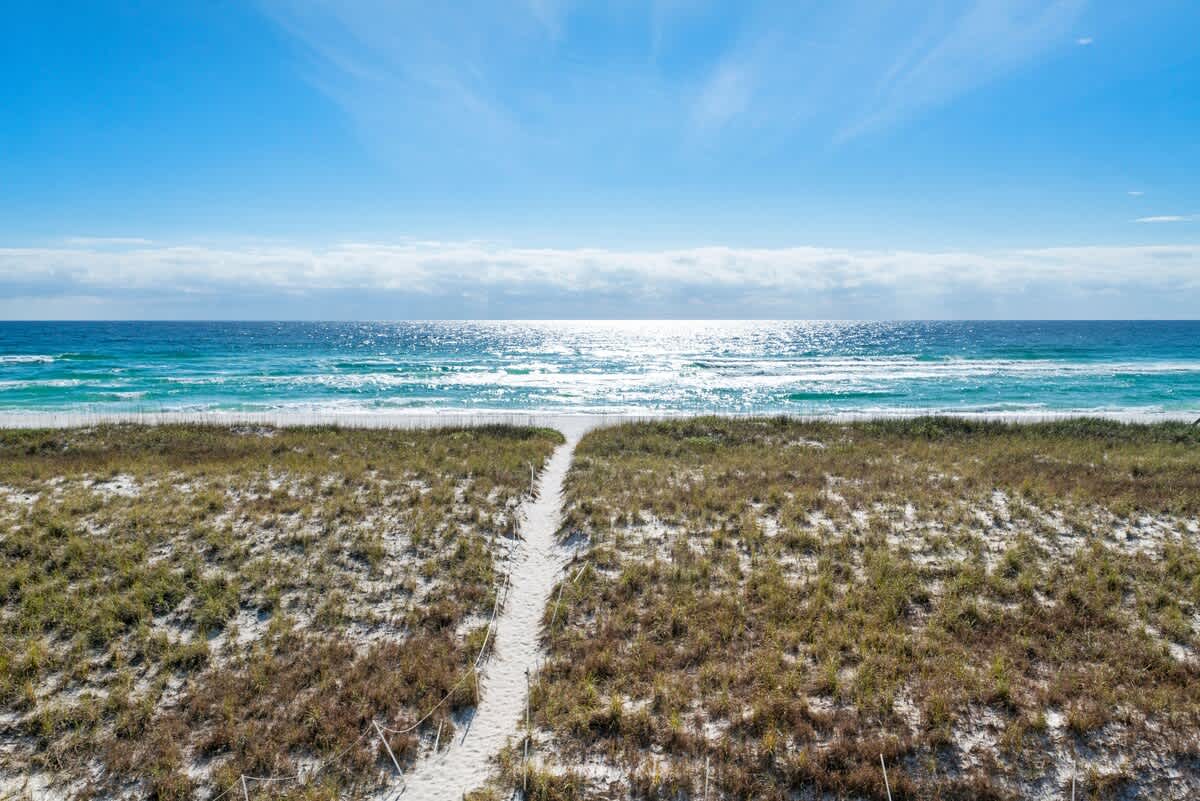 A pristine sandy path winds through lush green and brown grassy dunes, leading directly to the sparkling turquoise ocean under a bright blue sky with scattered clouds. The sun glints off the water, creating a shimmering horizon.