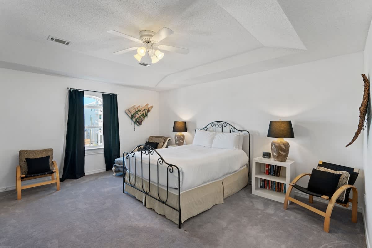 Spacious bedroom with light gray carpet, a black wrought iron queen bed, and two wooden chairs with black cushions near a window with dark curtains. A ceiling fan with lights provides illumination and air circulation.