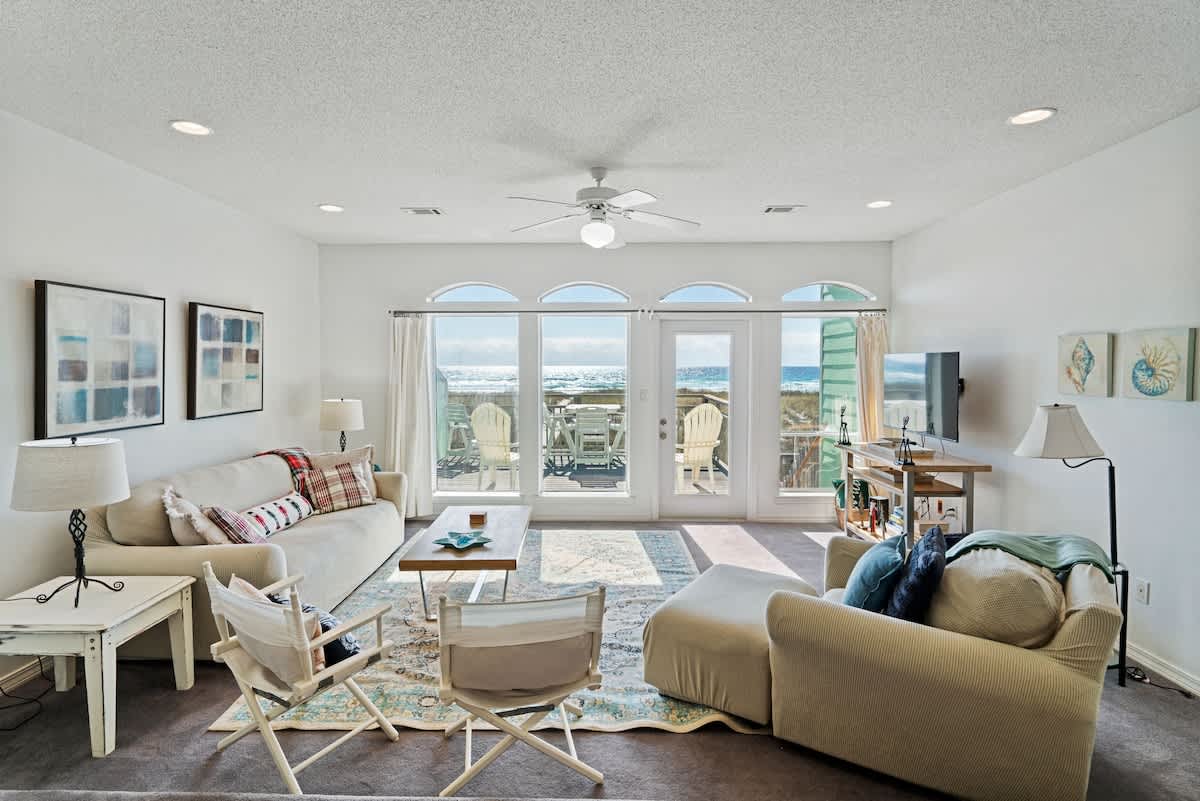 Spacious living room with a light-colored sofa and armchair, a wooden coffee table on a blue and beige patterned rug, and large windows providing a stunning ocean view.