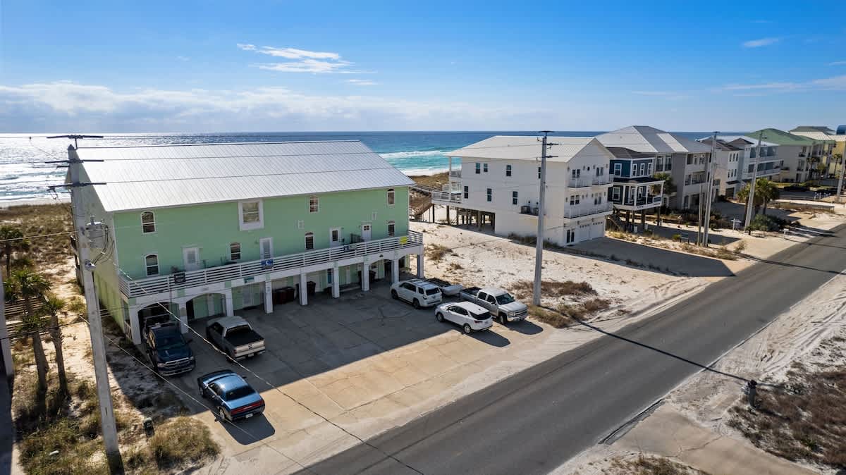 An aerial view showing the front of the light green townhouse with a parking lot, adjacent to a road, with the blue ocean visible in the background under a partly cloudy sky.
