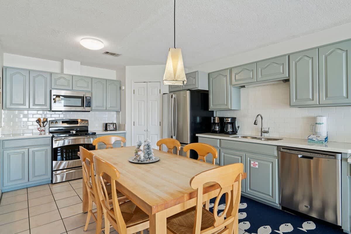 Well-equipped kitchen featuring light blue-gray cabinetry, white countertops, stainless steel appliances, and a large wooden dining table with chairs, all set on white tiled flooring.