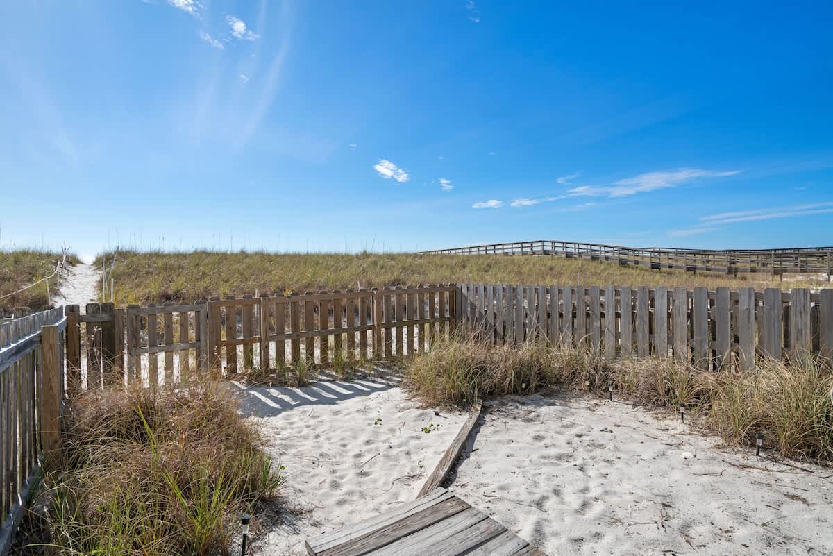 A private sandy yard area is enclosed by a low, weathered wooden picket fence, featuring sparse green and brown grasses. A wooden walkway leads from the foreground towards distant grassy dunes and a boardwalk under a clear blue sky.
