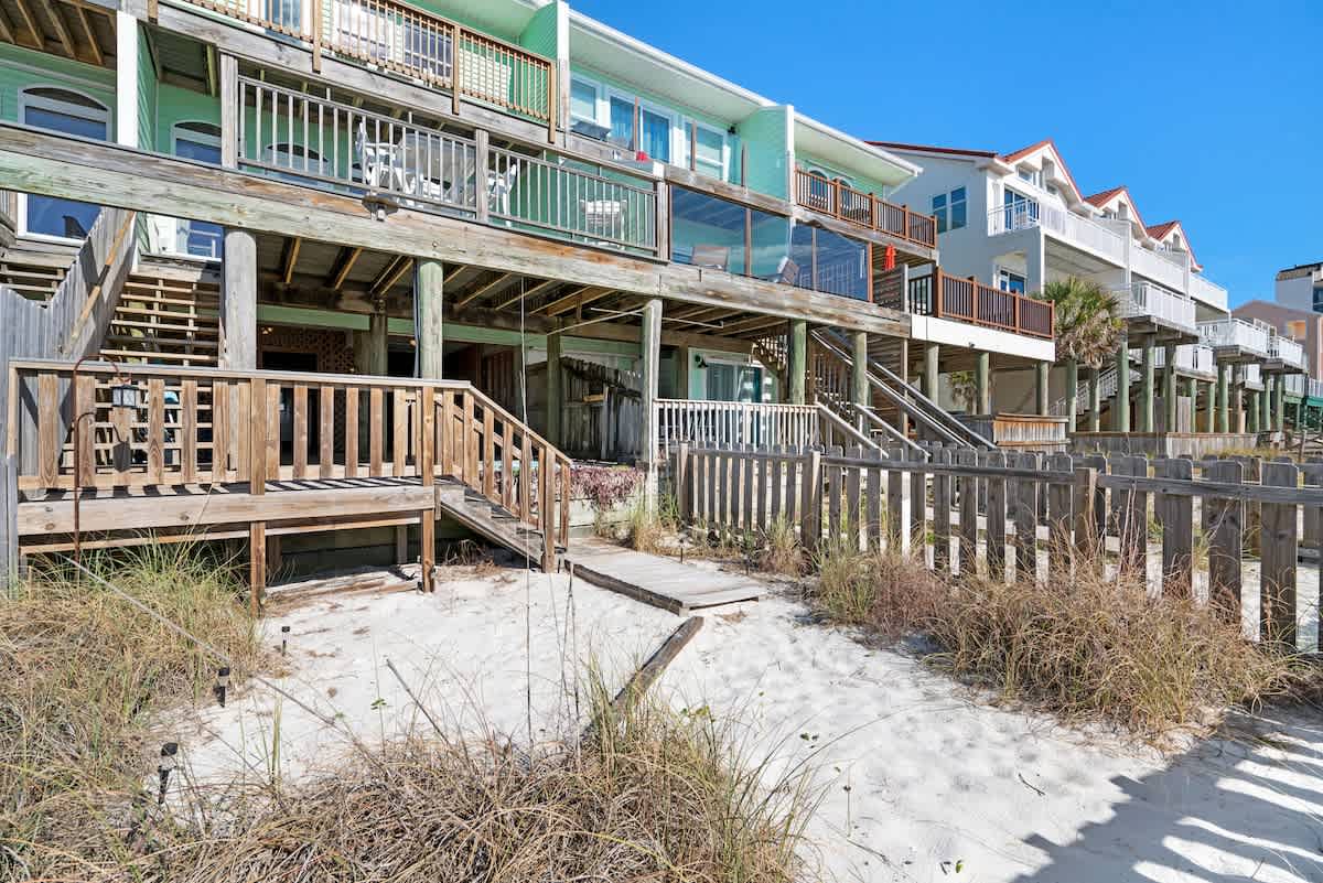 A row of multi-level beach houses with wooden decks and stairs, leading down to a sandy area with a wooden fence and direct beach access, under a bright blue sky.