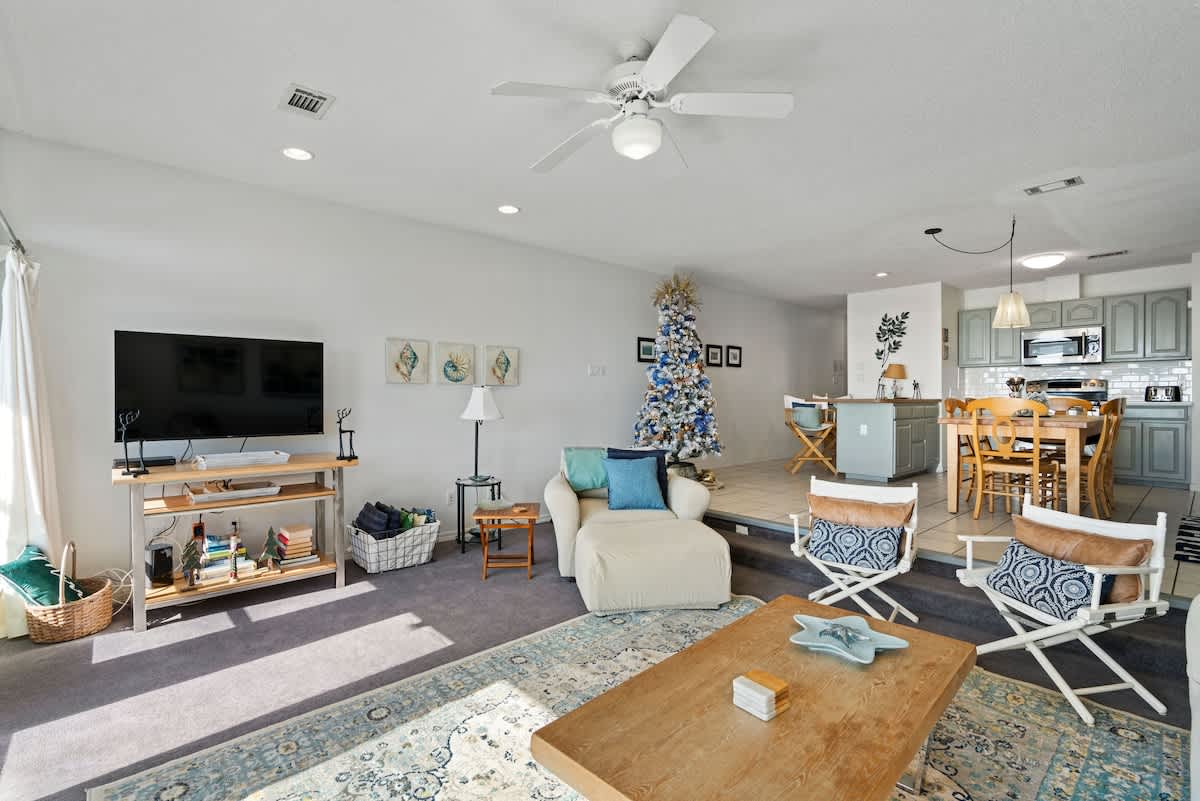 Open-concept living area with a light-colored armchair and sofa, a wooden coffee table, and a TV stand, leading into a dining area and kitchen, with a decorated Christmas tree in the background.