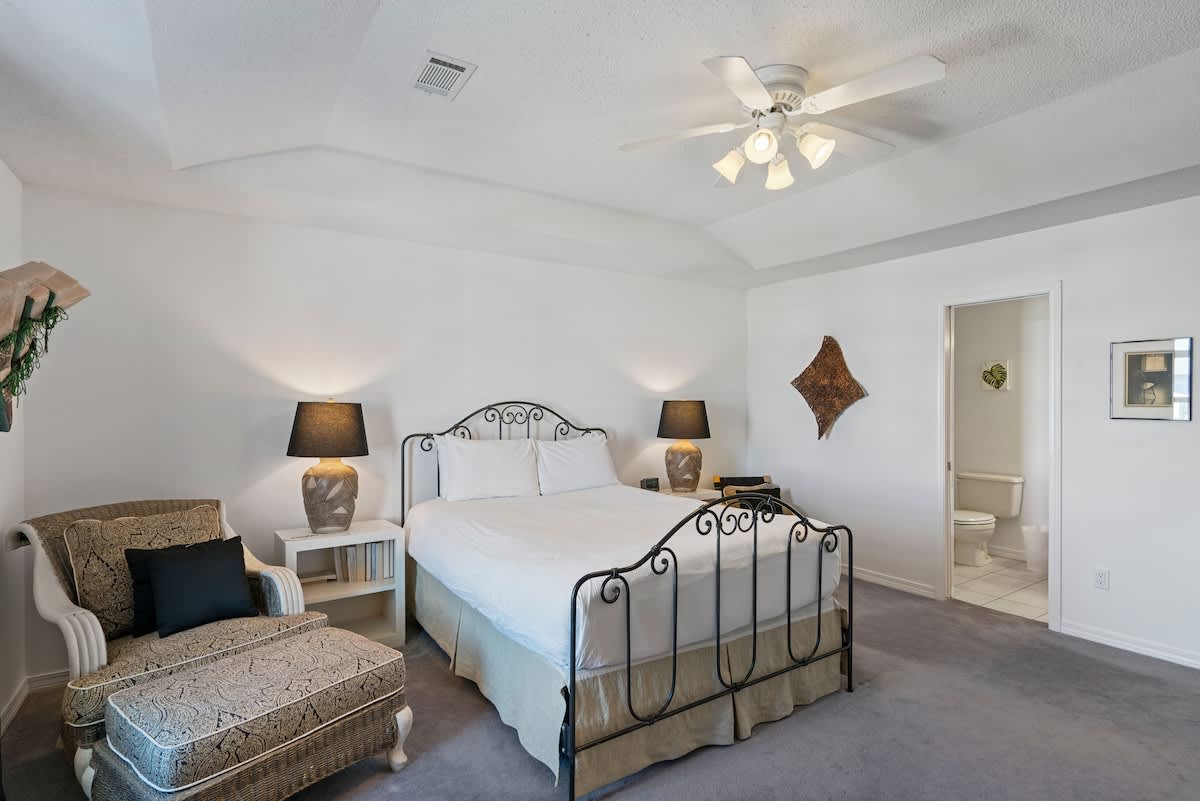 Bright bedroom with light gray carpet, a black wrought iron queen bed, and a patterned armchair with an ottoman. An open doorway leads to an en-suite bathroom, enhancing privacy and convenience.