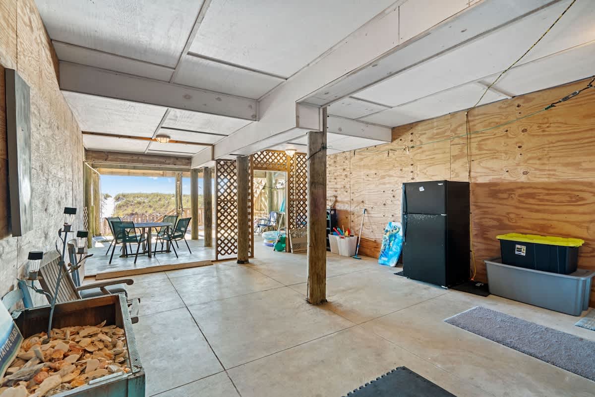 Spacious covered outdoor area under the house with a concrete floor, exposed wooden beams, and support pillars. It features a black refrigerator, various beach gear, and a view towards the beach.