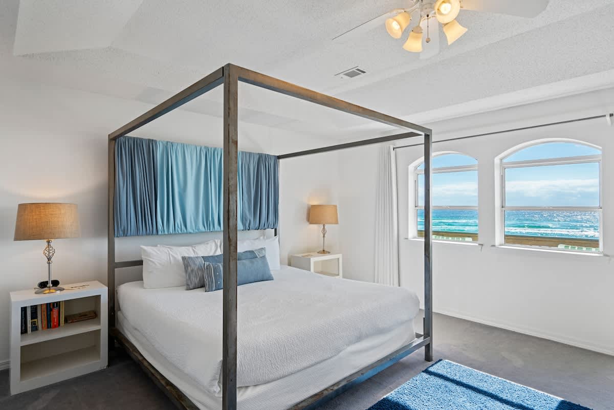 King-sized metal canopy bed with white bedding and blue pillows, flanked by white nightstands, with arched windows providing stunning ocean views.