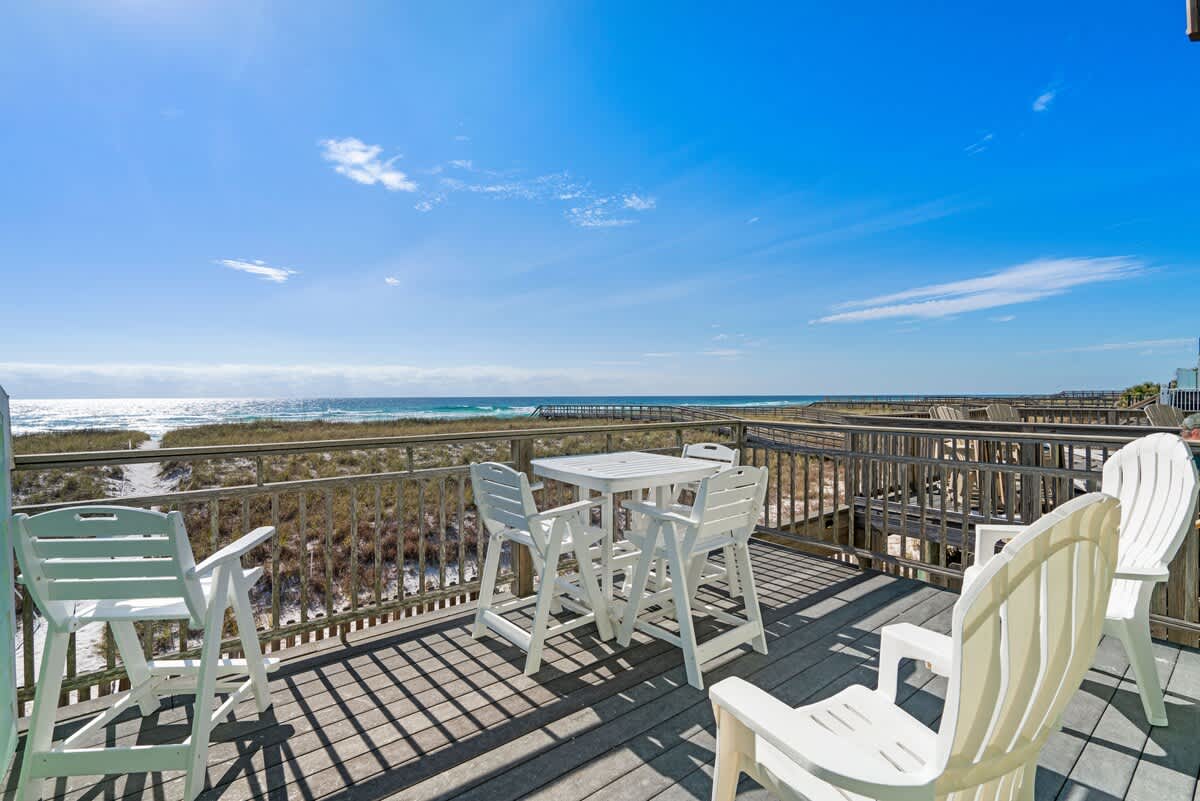 A spacious wooden deck with a white plastic dining set and several chairs, offering panoramic views of the sandy beach, dunes, and the sparkling blue ocean under a clear sky.