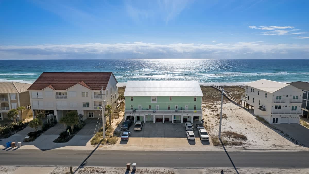 A higher aerial view of the light green townhouse and surrounding properties, with the vast blue ocean and white sand beach in the background, under a bright blue sky.
