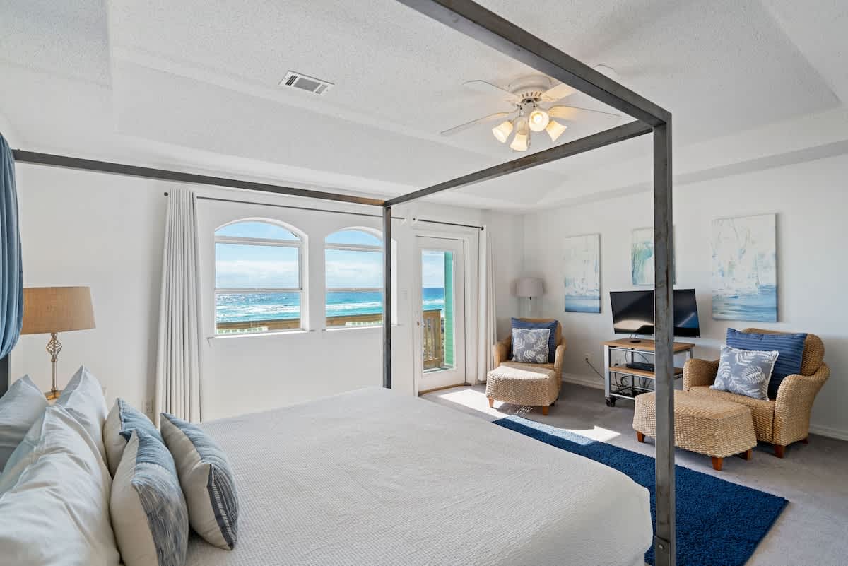 Master bedroom with a king canopy bed, a cozy sitting area with wicker chairs and ottomans, and a glass door leading to a balcony with ocean views.